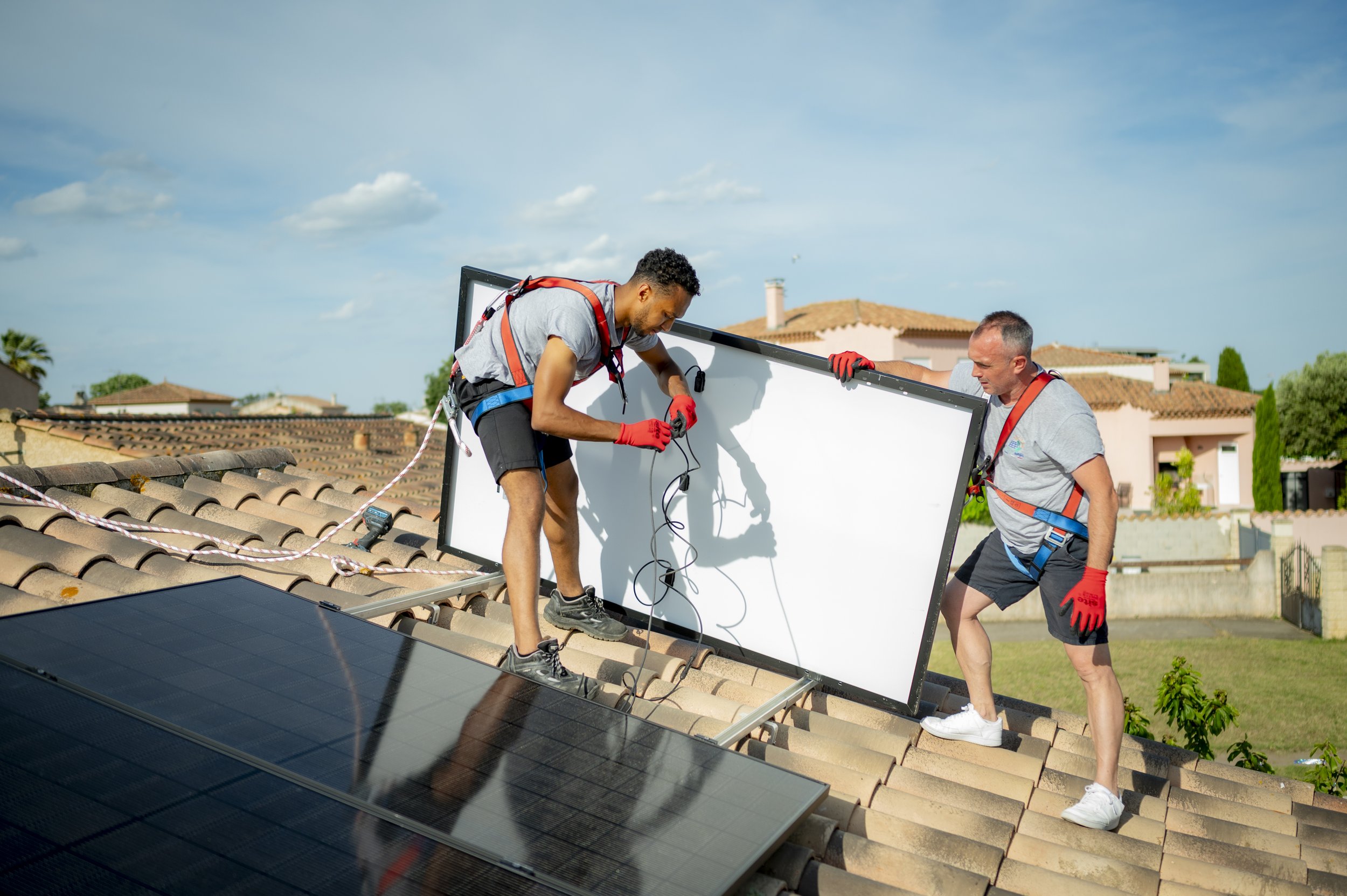 Deux techniciens installent des panneaux solaires sur un toit en tuiles, ensoleillé avec quelques nuages dans le ciel.