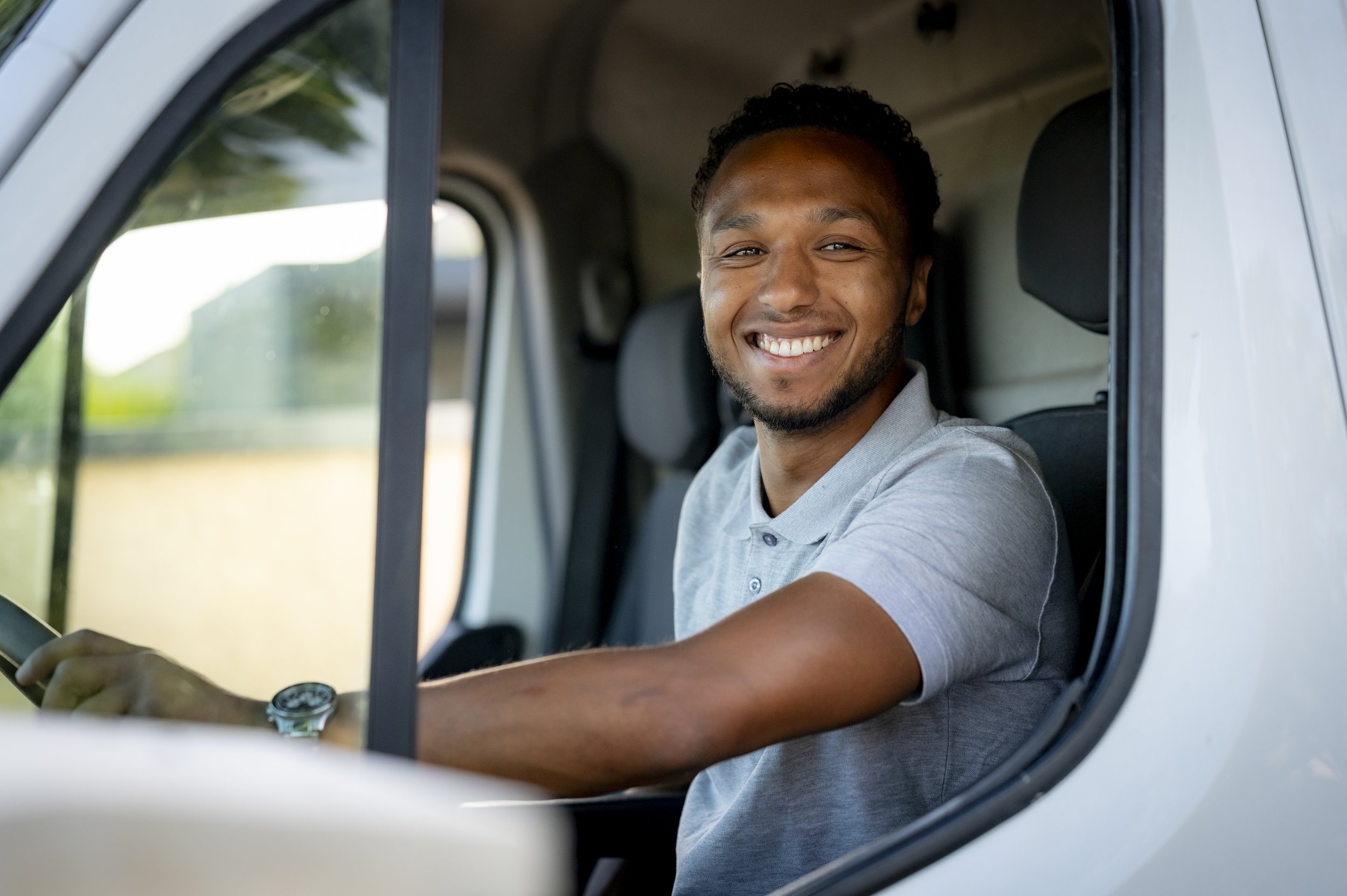 Homme souriant assis au volant d'un véhicule blanc, portant un polo gris.
