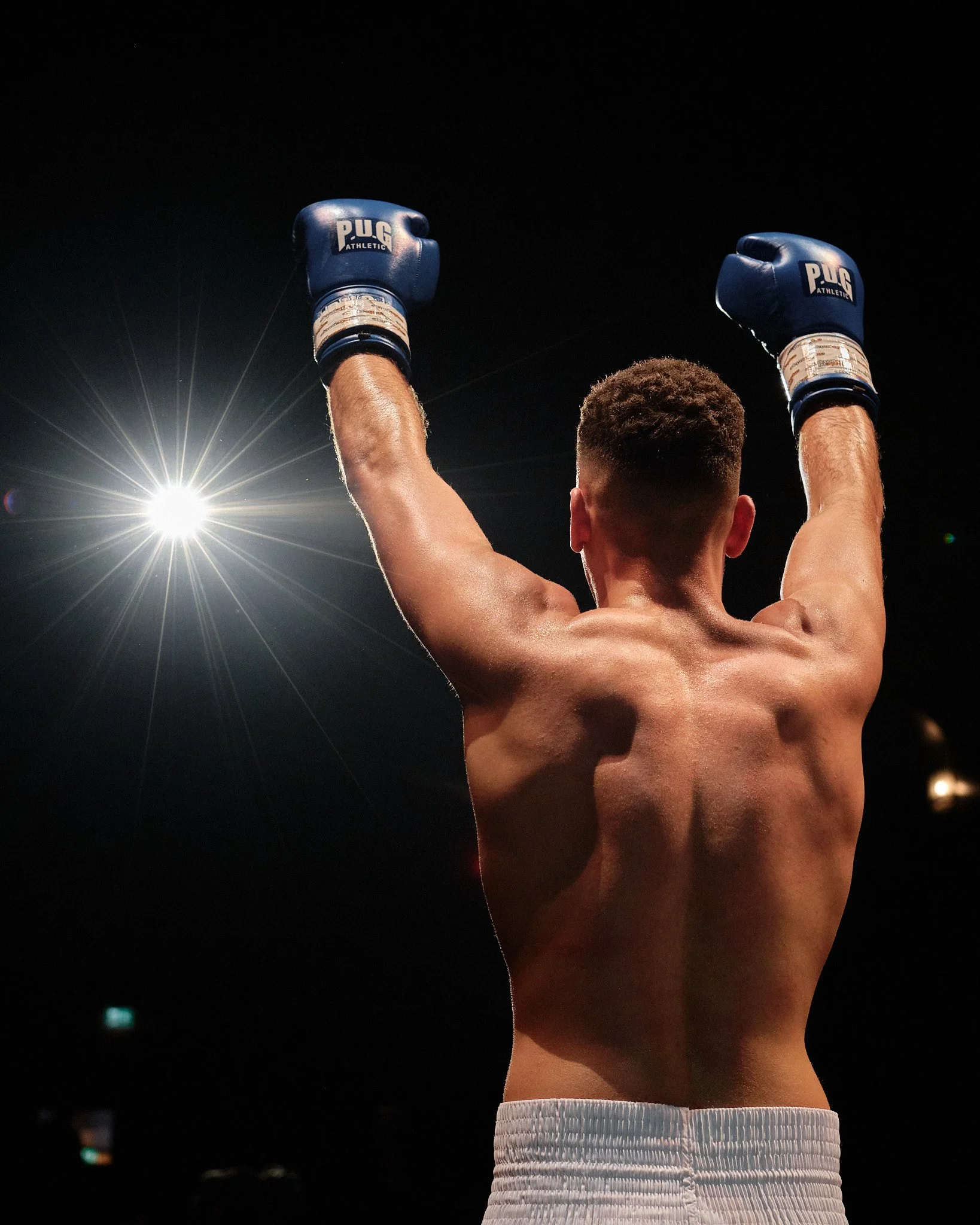 Boxer wearing blue gloves with raised arms, illuminated by a bright light in a dark environment.