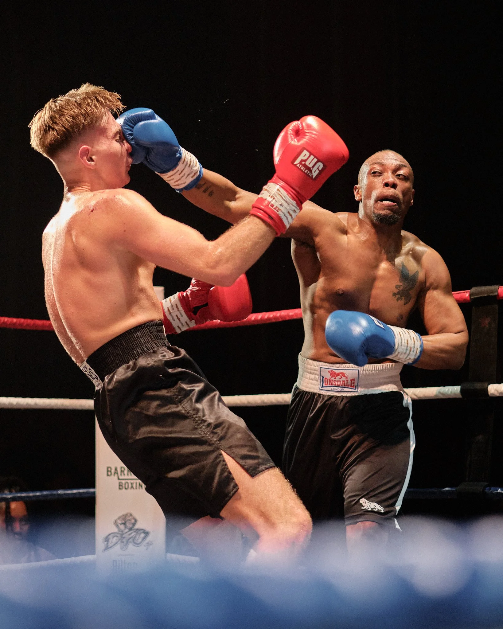 Two male boxers in a ring during a match, one throwing a punch and the other blocking, both wearing boxing gloves and shorts.