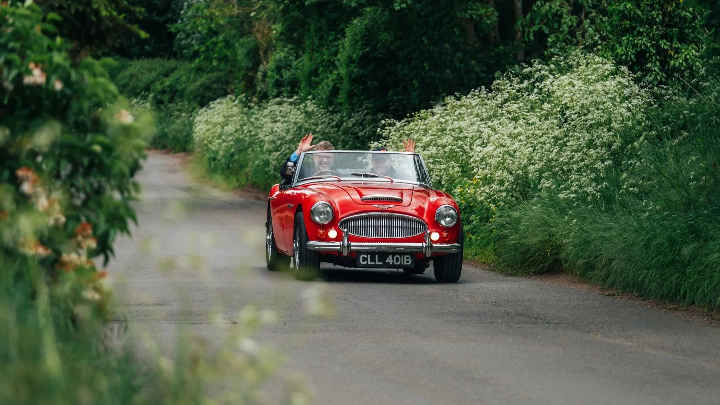 A red vintage convertible car with two people waving, driving along a narrow country road surrounded by green bushes and trees.