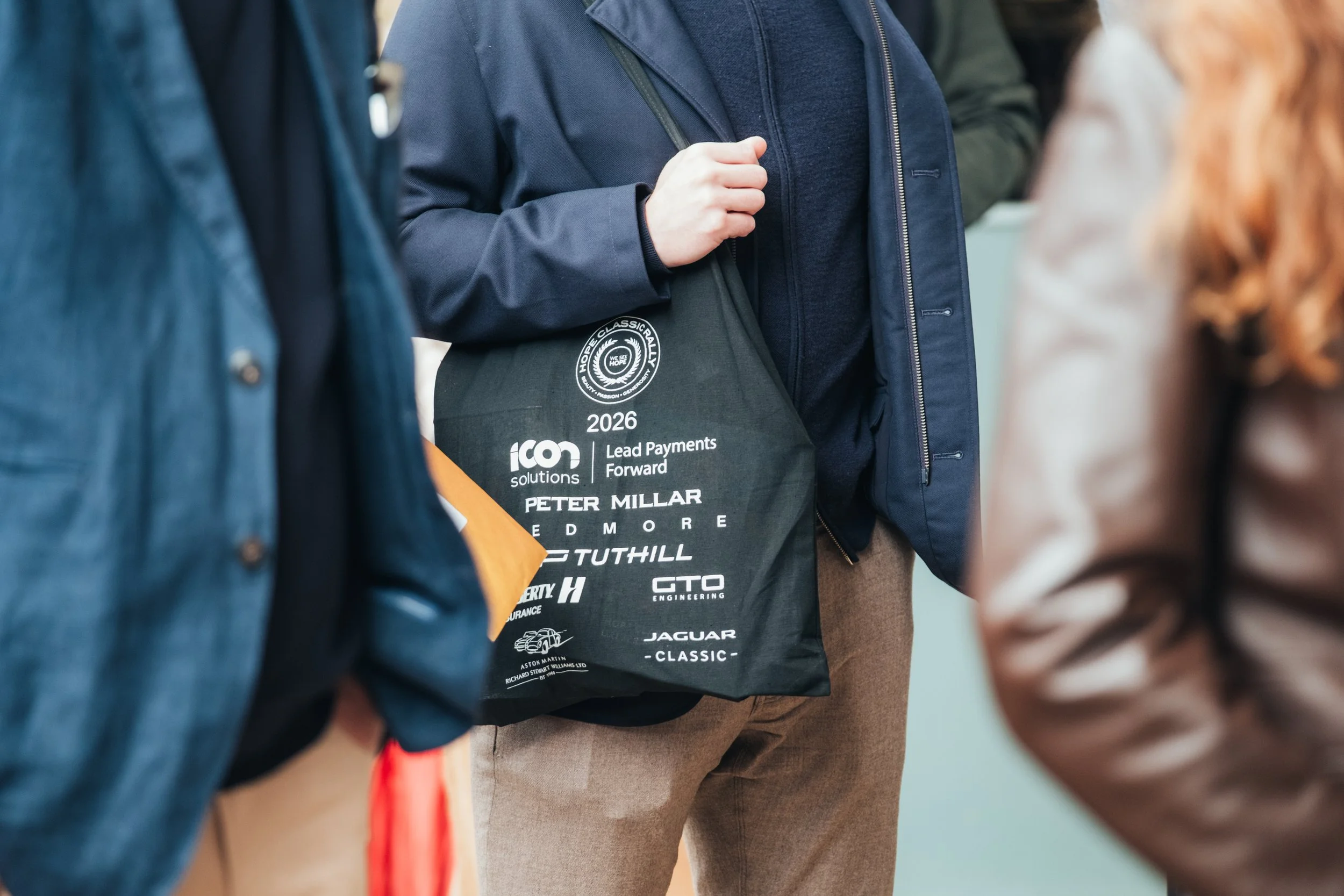 Close-up view of a person wearing a navy blue jacket and holding a black tote bag with white text and logos, surrounded by other people.