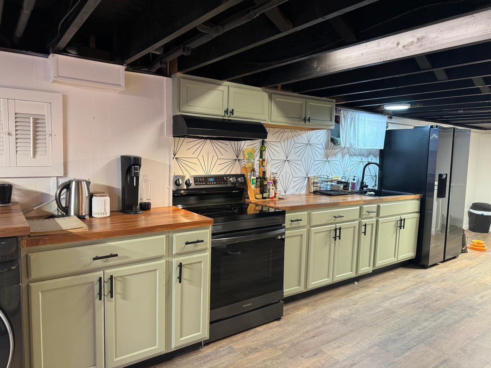Basement kitchen with light green cabinets, black stove, and wooden countertop. A black refrigerator is visible, along with a coffee maker, tea kettle, and coffee canister on the counter. The backsplash features a geometric pattern, and the ceiling h