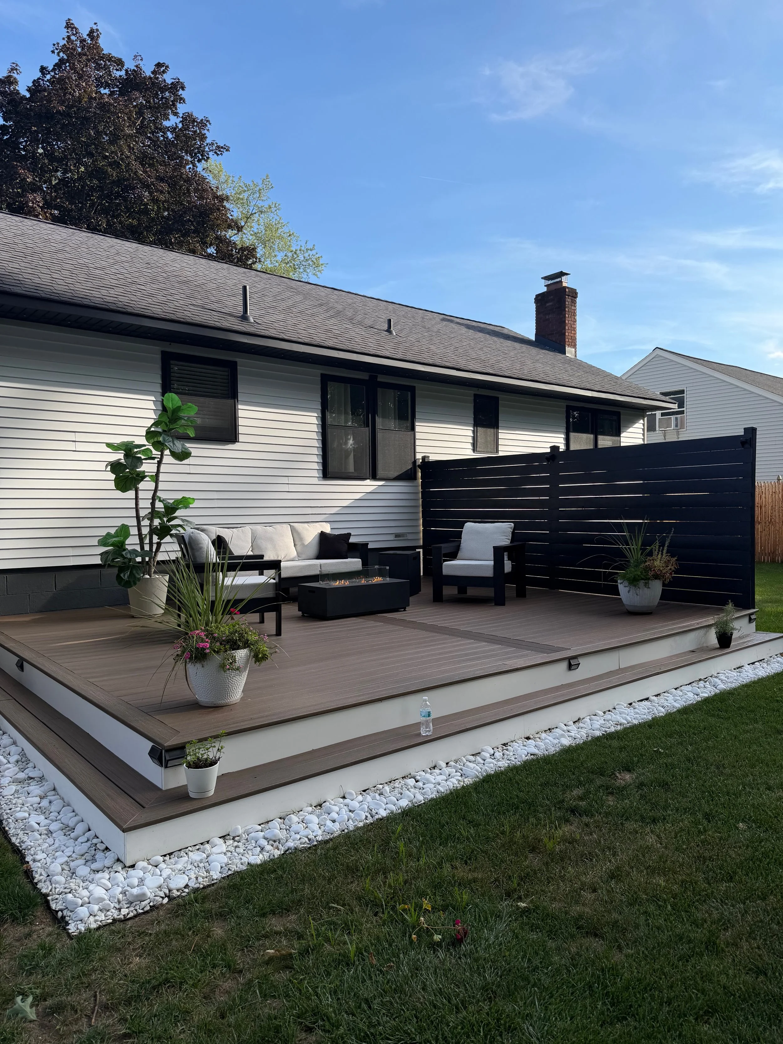 Completed Trex composite deck featuring a wraparound step, new white siding, and black new-construction windows for a clean, modern exterior. The custom privacy wall was painted black to coordinate with the windows and exterior details, creating a co