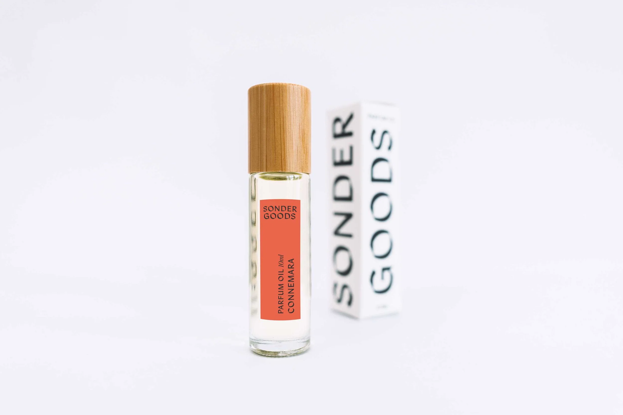 A clear glass perfume bottle with a wooden cap, labeled SONDER GOODS, containing 10ml of orange oil labeled CONNEMARA, stands next to a white box with black text reading SONDER GOODS on a white background.
