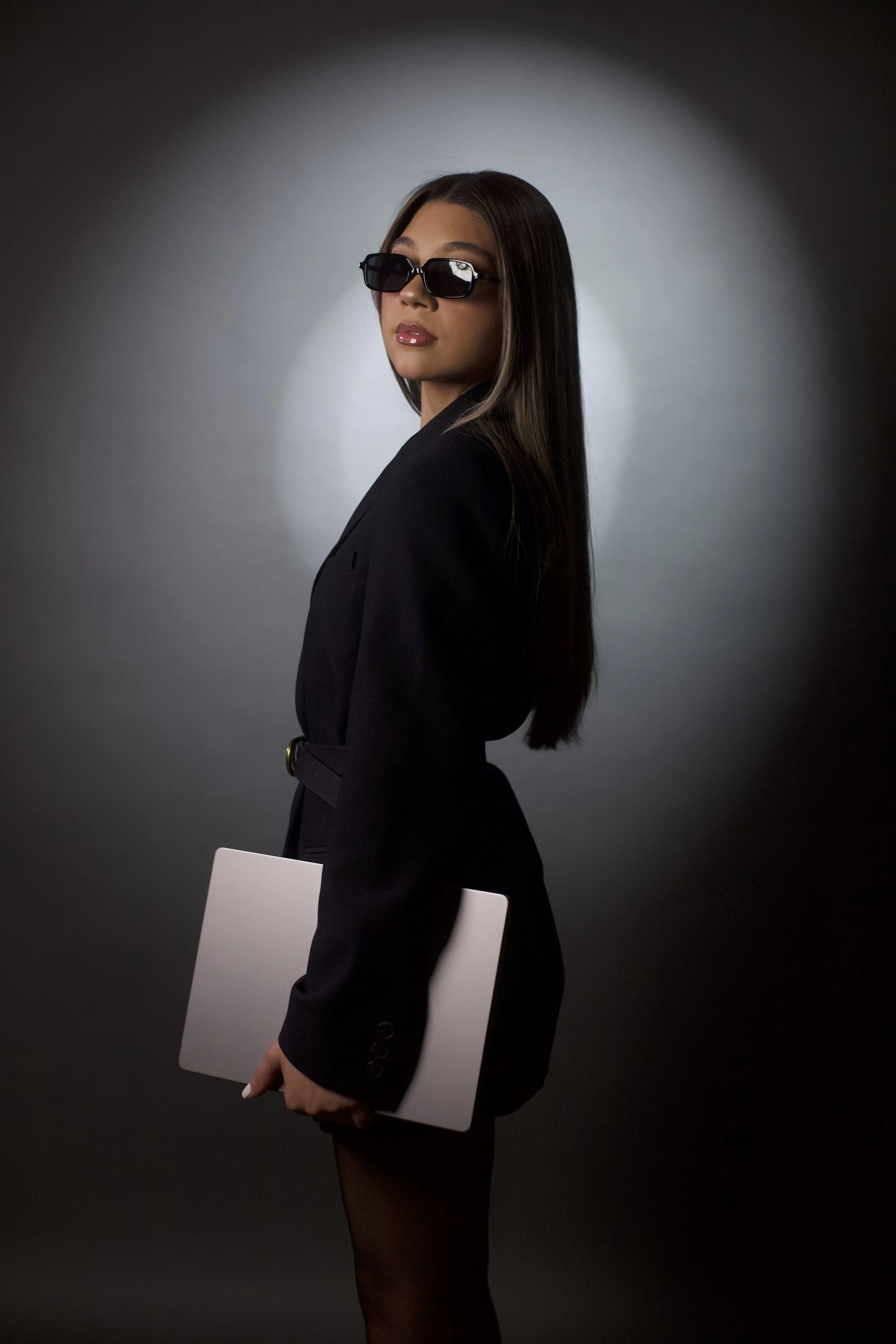 A stylish woman in a black suit with sunglasses, holding a closed laptop, standing against a grey background with a spotlight effect.