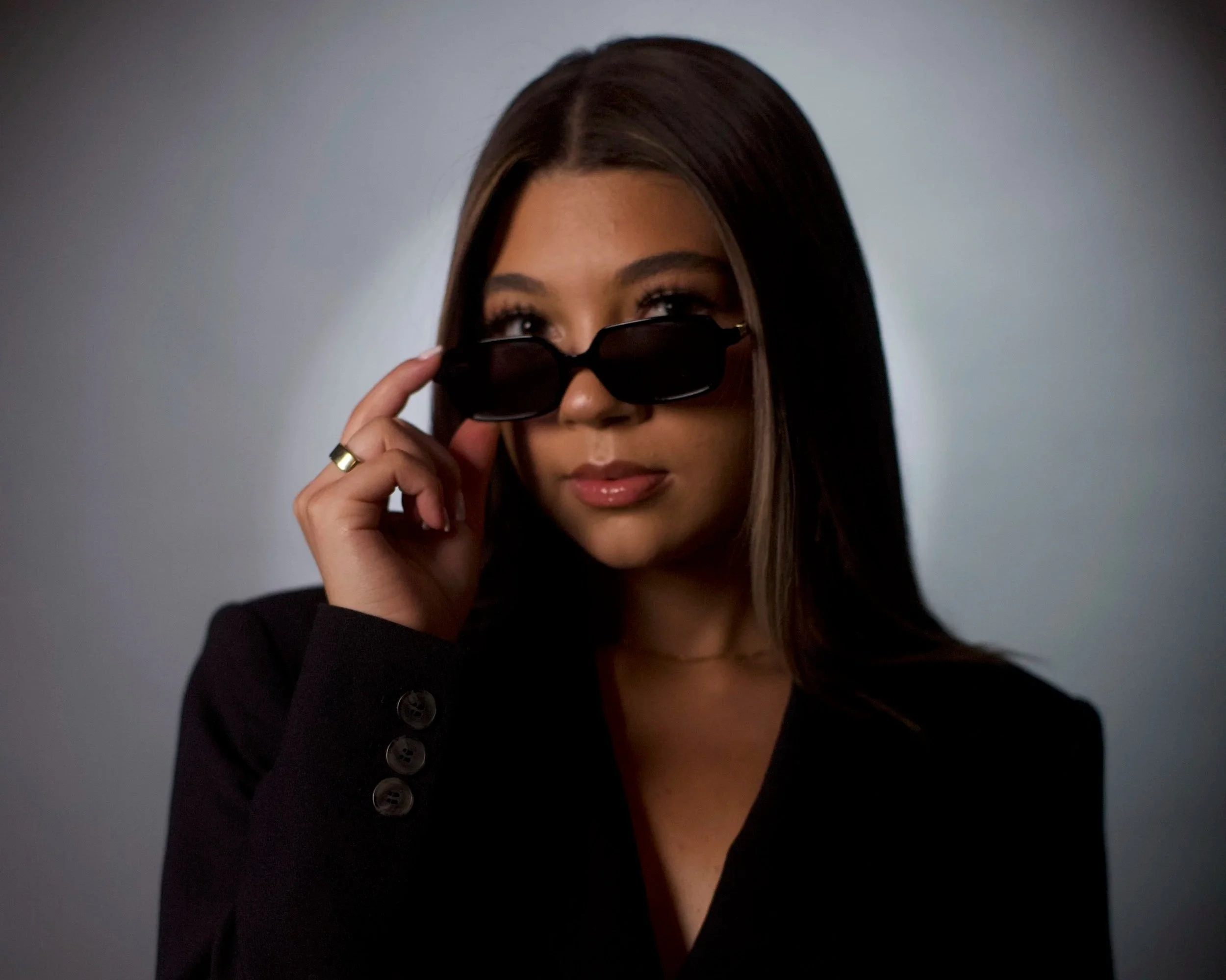 A woman with long dark hair in a black blazer, holding sunglasses and looking over them with a confident expression, against a plain gray background.