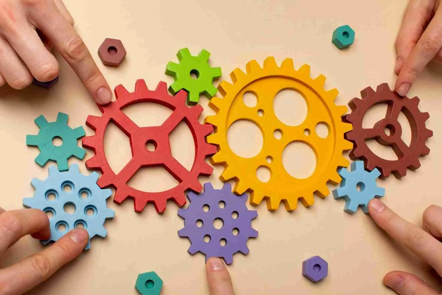 Colorful interlocking gears and hexagonal nuts with hands pointing at them on a beige background.