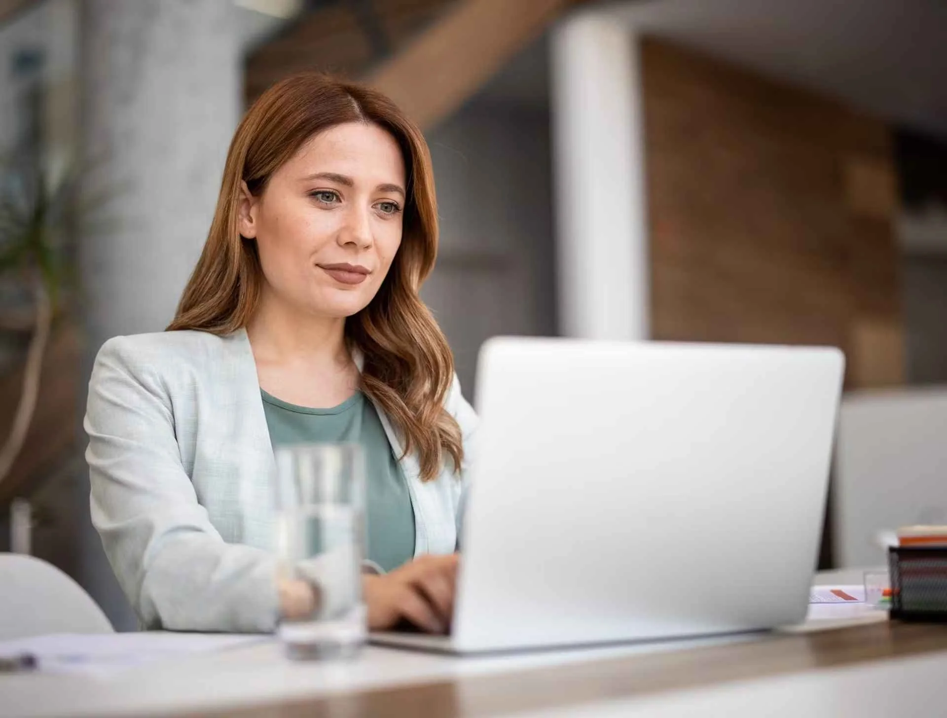 A woman with long, wavy hair working on a laptop at a desk in an office environment.