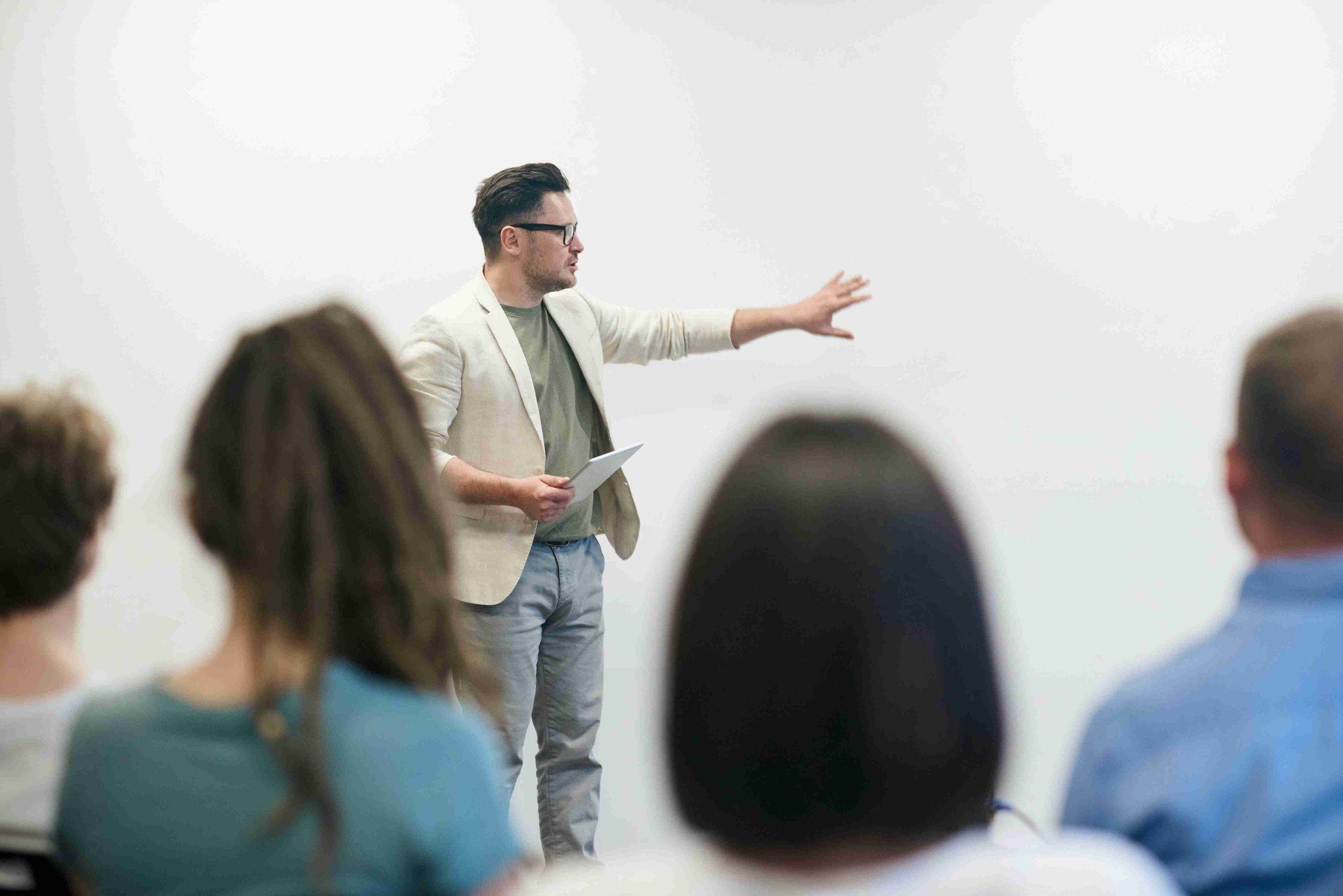 Man in light blazer giving a presentation to a group of people in a room with a white wall.