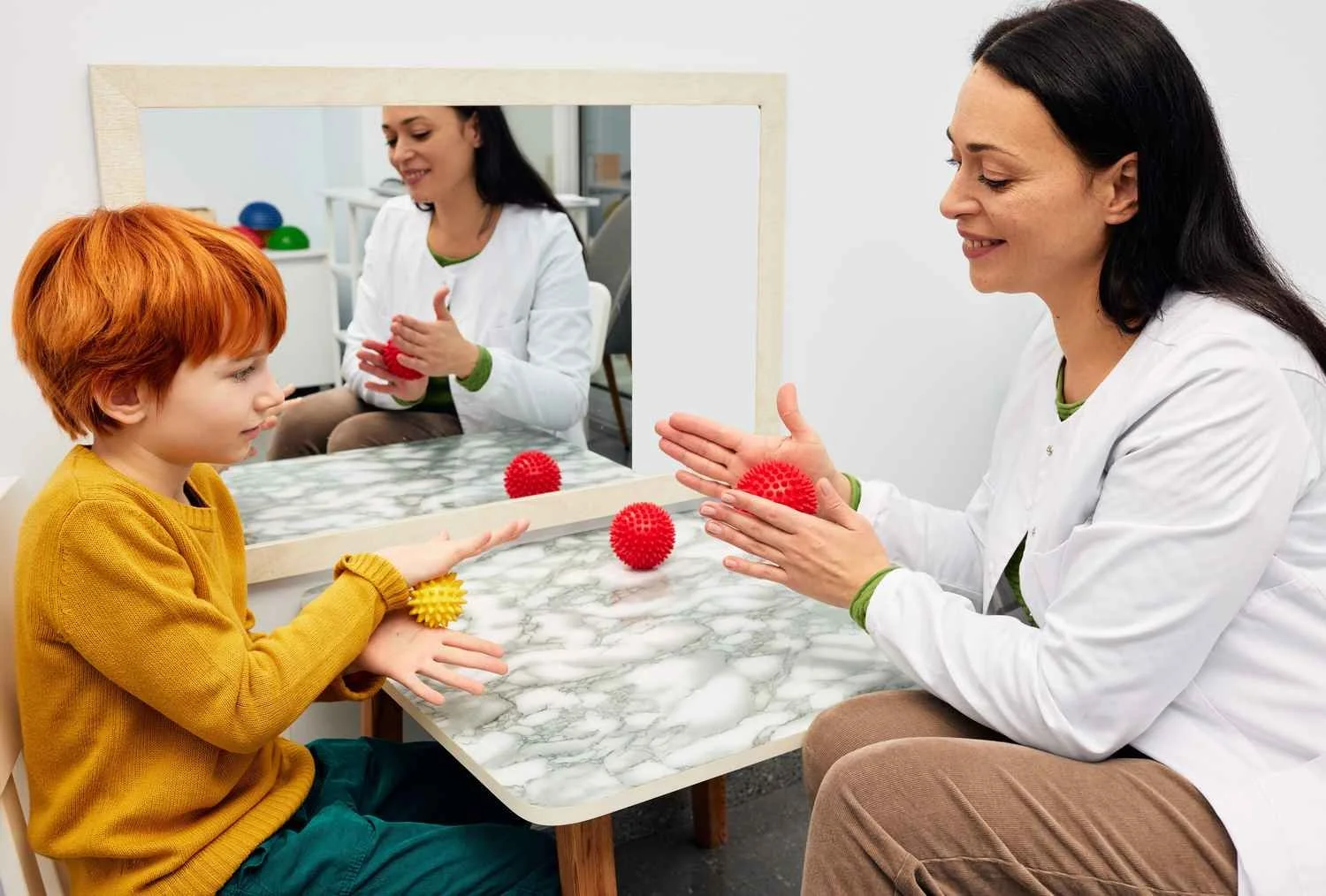A young boy and a female doctor are sitting at a table with a mirror in front of them. The boy is holding a yellow spiky ball, and the woman is holding three red spiky balls. The woman is demonstrating something to the boy with the balls, and her reflection is visible in the mirror.