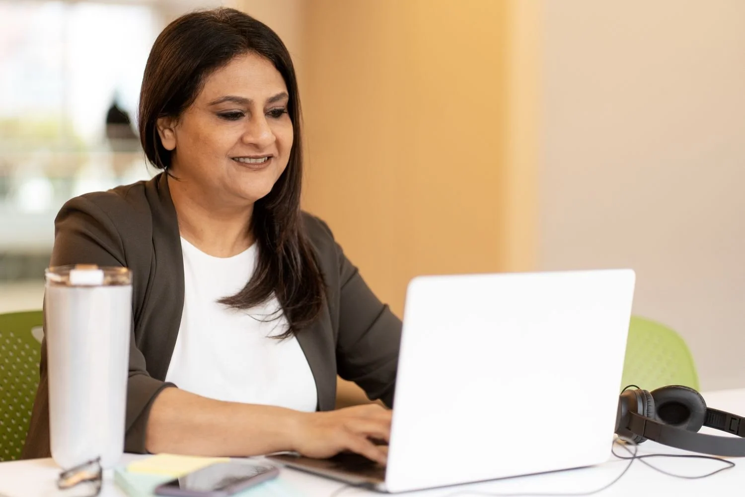 A woman with dark hair working on a laptop at a desk, with headphones and a water bottle nearby.