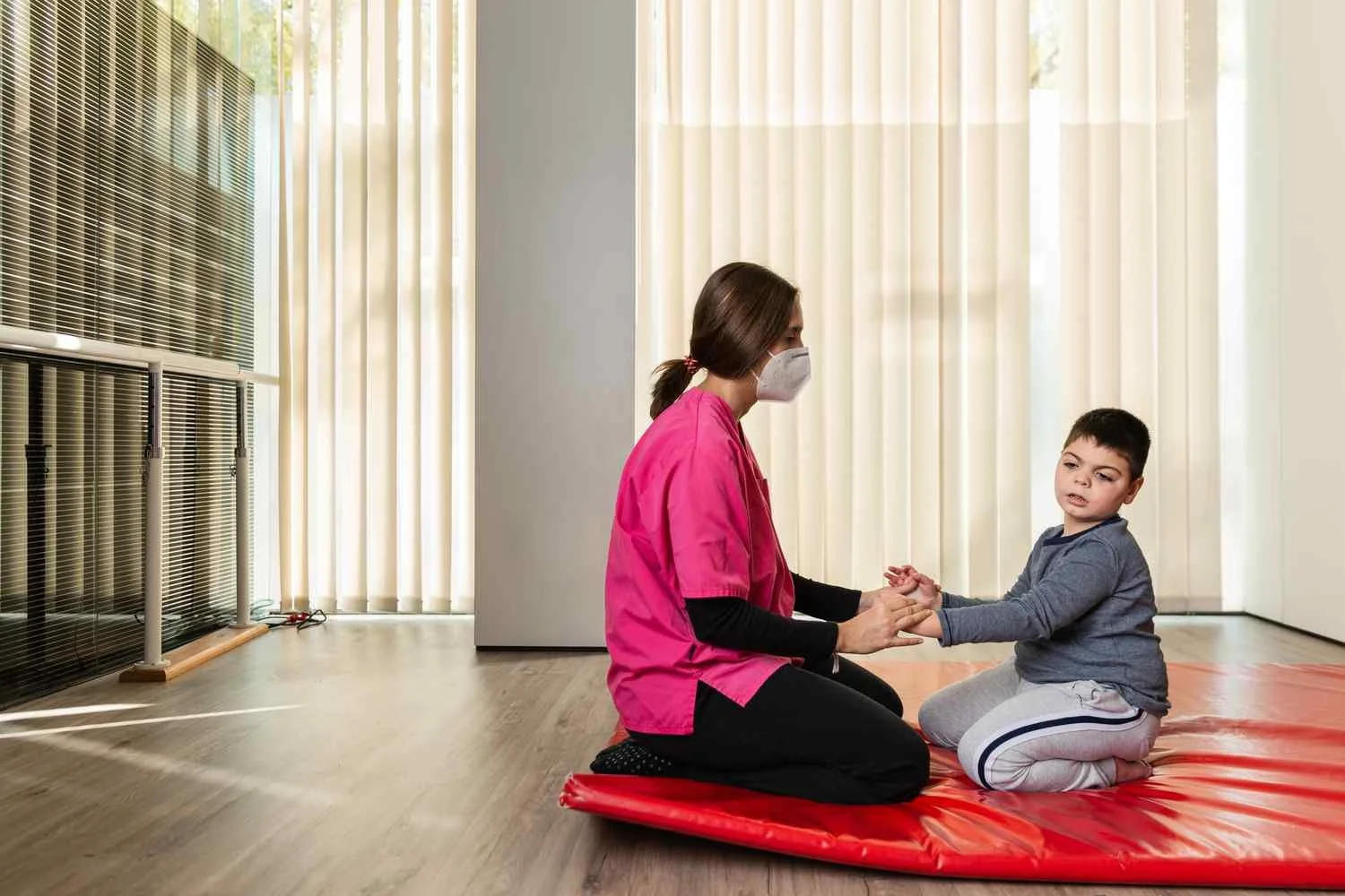 A therapist and young boy sitting on a red mat in a room with sunlight streaming through vertical blinds. The therapist wears a pink uniform and a face mask, holding the child's hands, who is looking at her with a serious expression.