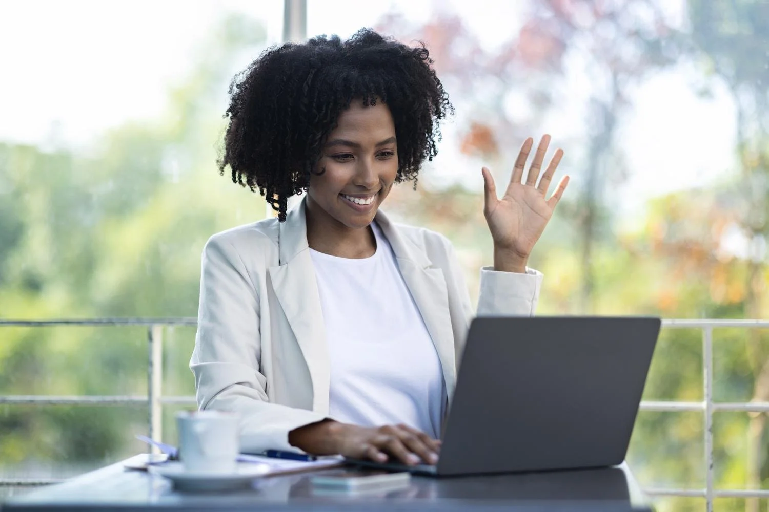 Smiling woman with curly hair waving while sitting at a table and working on a laptop.
