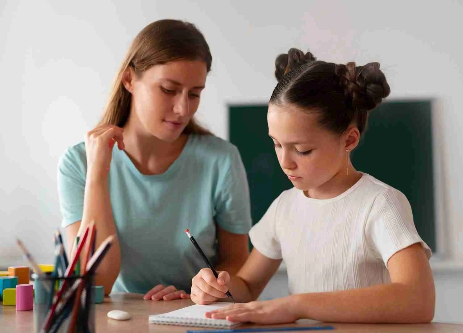 Teacher and student at a desk, with the student writing and teacher observing.