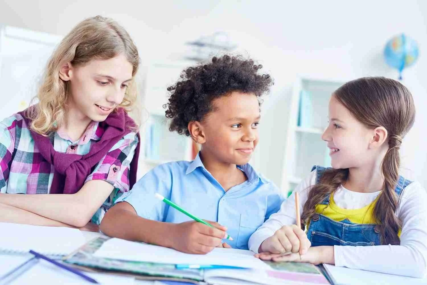 Three children sitting at a table, smiling and working together on a project, with books and papers in front of them.