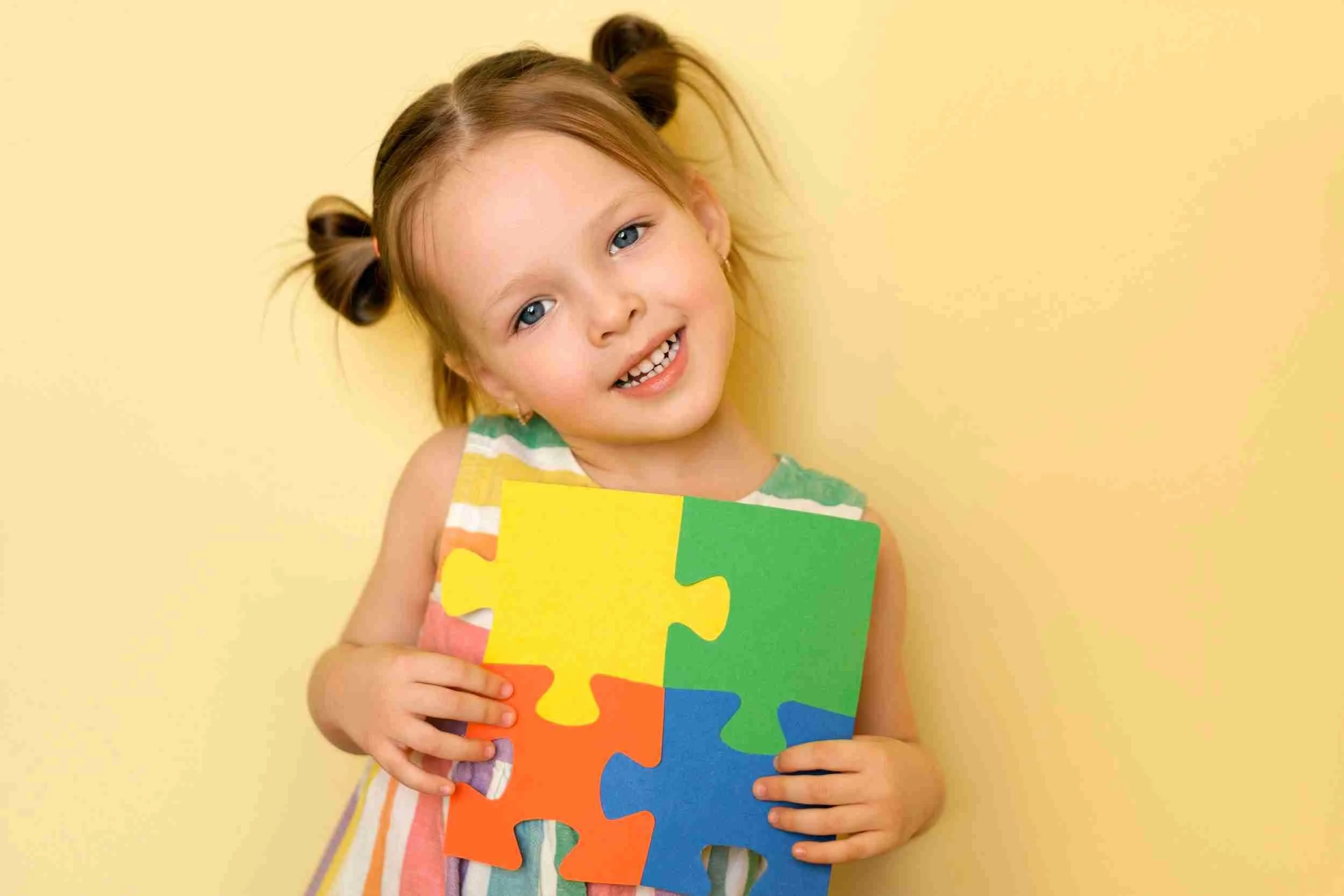 Young girl holding colorful puzzle pieces against yellow background.