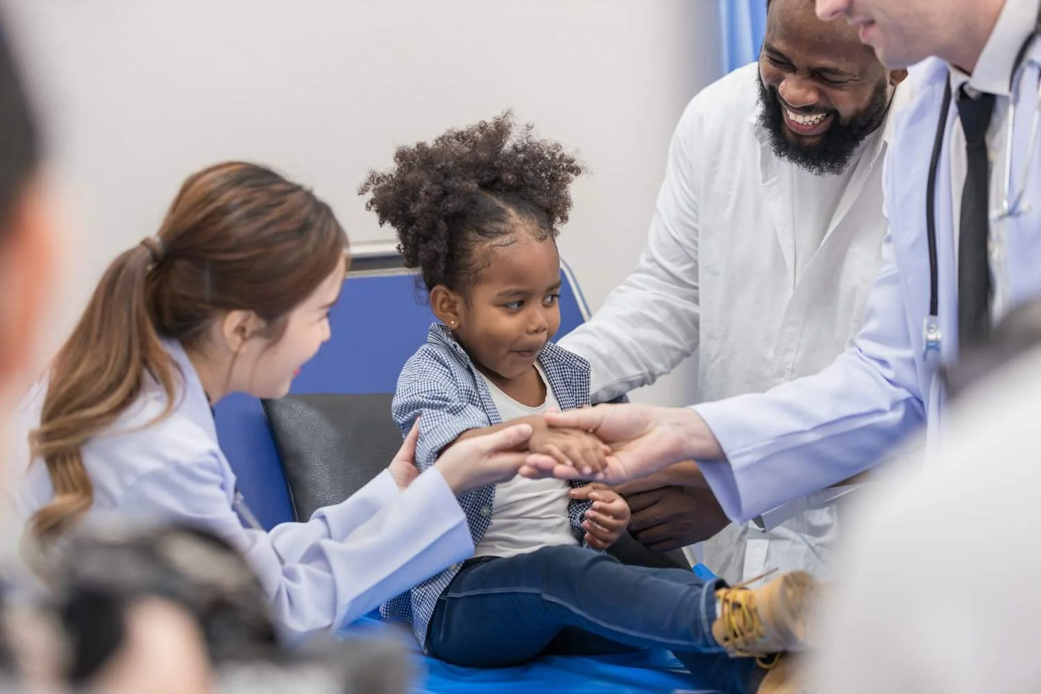 Young girl in a checkered shirt sitting on a doctor's examination table, shaking hands with a medical professional, while a doctor and a man smile nearby in a medical office.