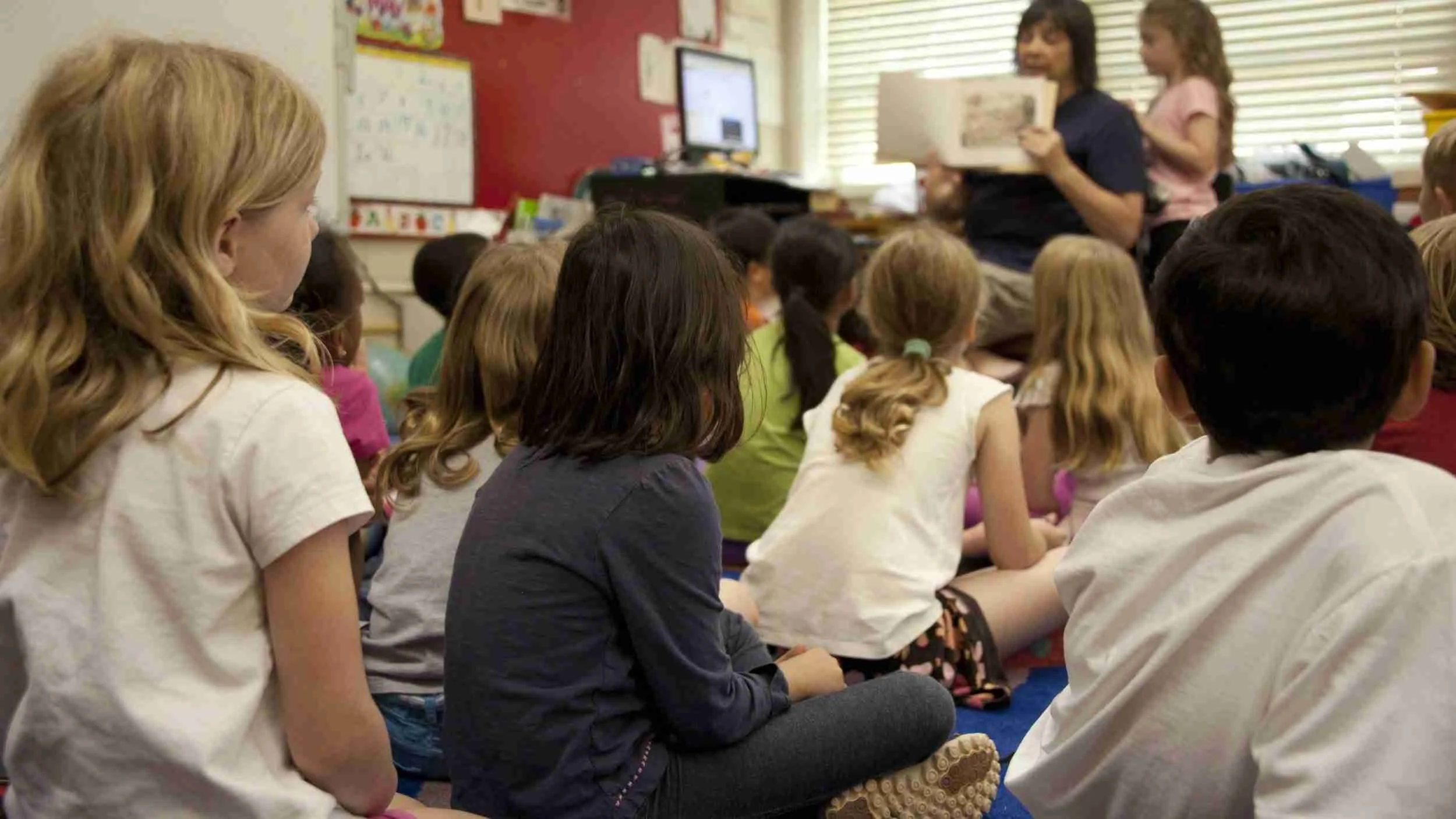 A group of young children sitting on the floor in a classroom, listening to a teacher read a book.