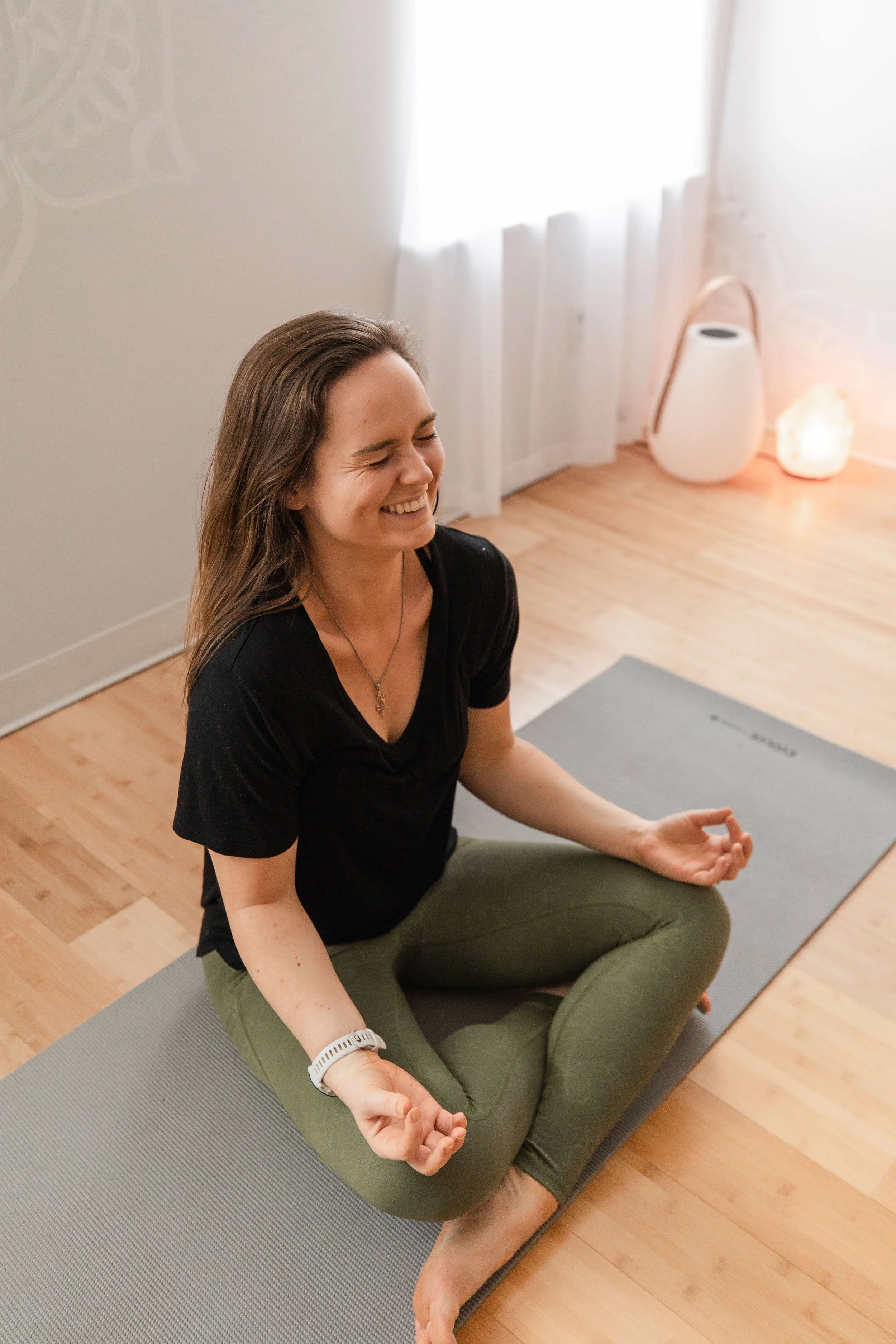 Tyler Mauler sitting cross-legged on a yoga mat in a bright room, smiling with her eyes closed as she practices meditation.