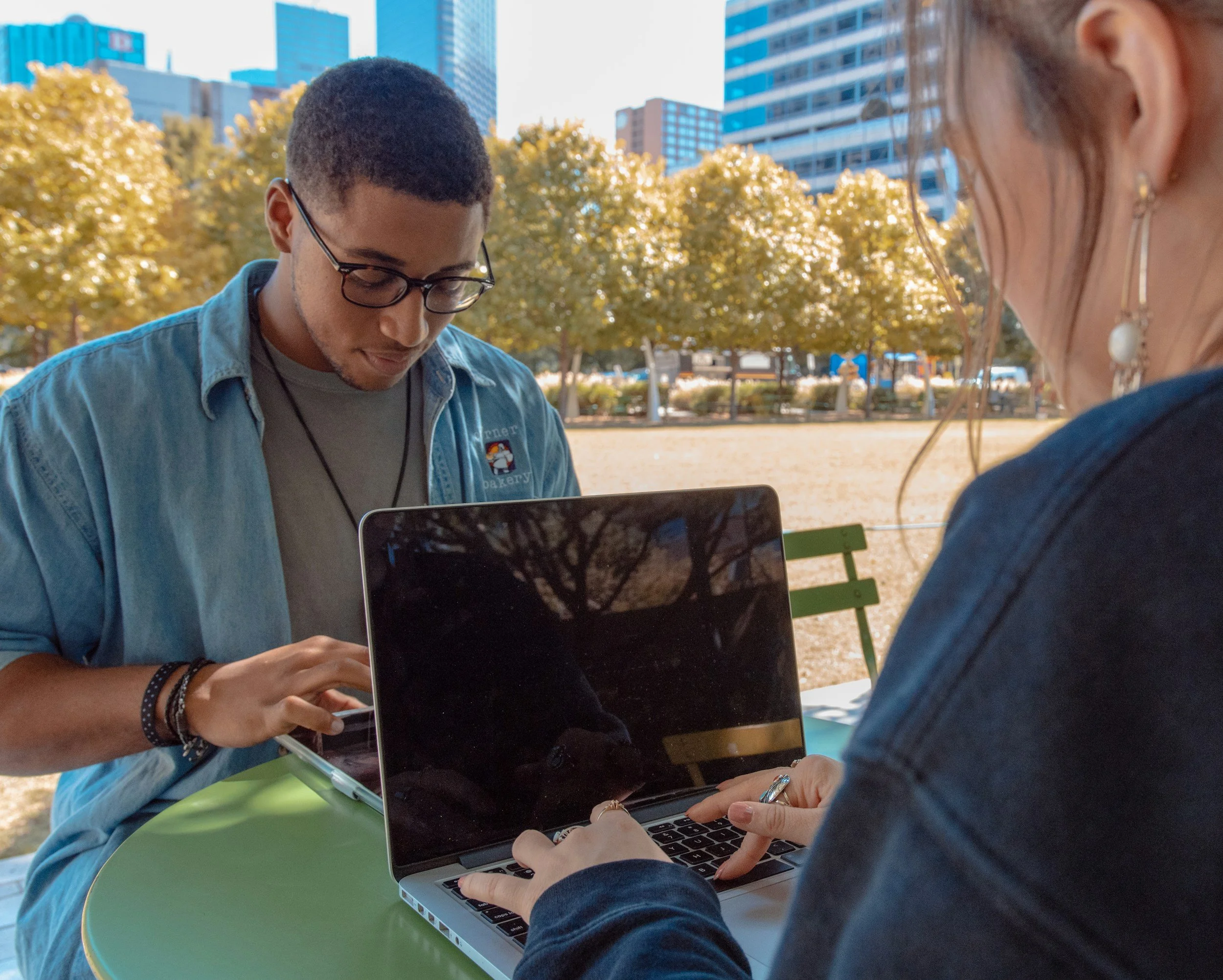 Two high school students sitting at a green table outdoors on a sunny day, working on laptops. There are trees with autumn-colored leaves and tall buildings in the background.