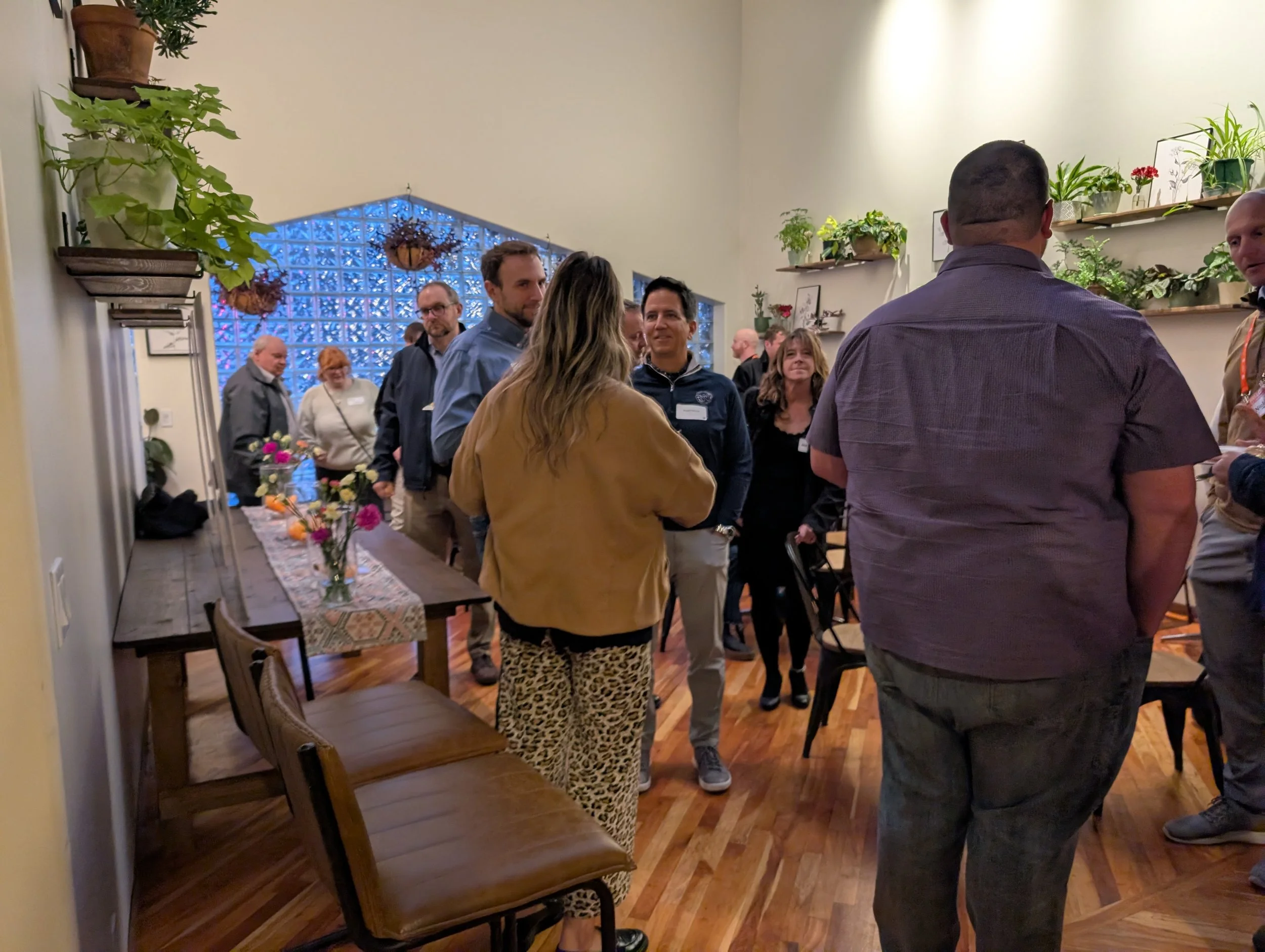 Group of people gathered in a room with wooden floors, some conversing and others standing near a photo display. There are wall-mounted shelves with potted plants, and a long table with a floral centerpiece and fruit. The room has a decorative glass block window and hanging plant baskets.