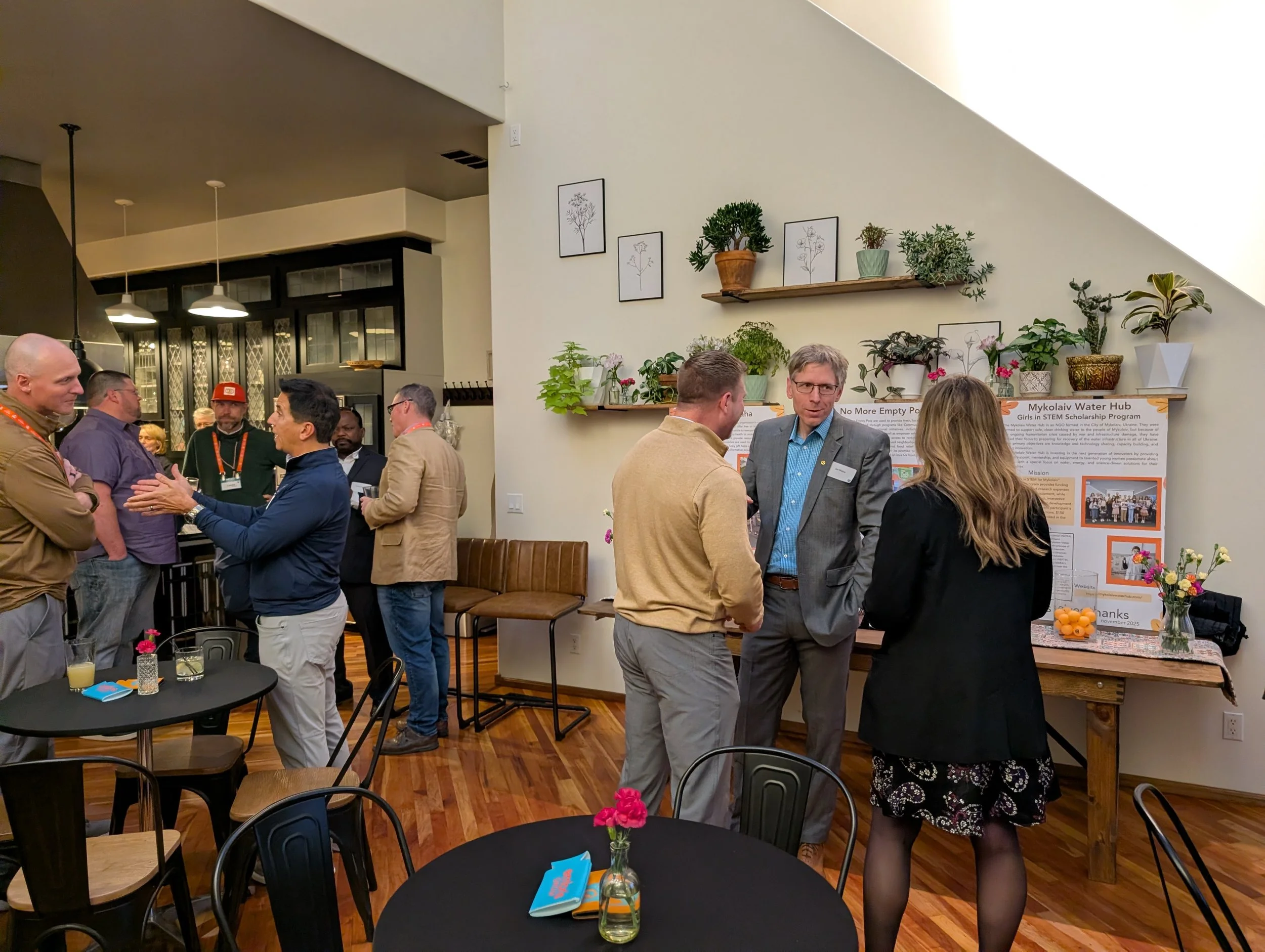 People engaged in conversations at a professional event, with a display board about a water hub project in the background and tables with flowers in the foreground.