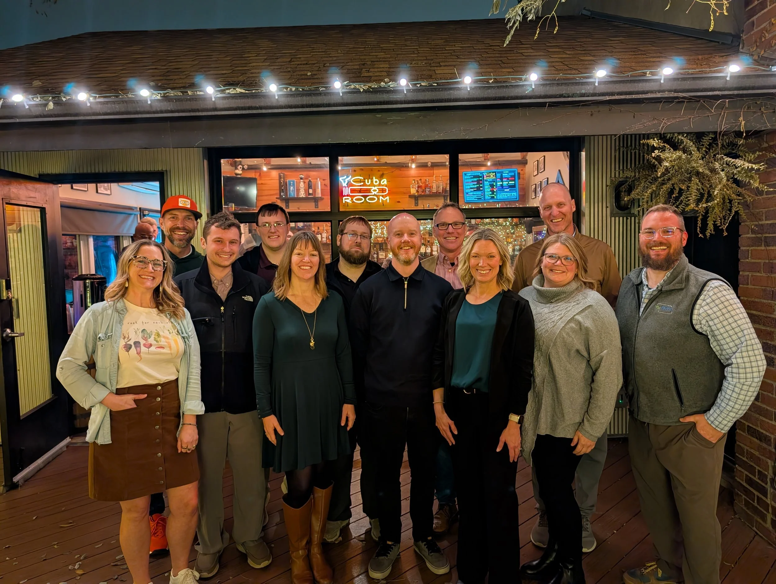 A group of 12 people smiling and posing for a photo outside a restaurant or bar called Cuba Room at night. The group is dressed casually, some wearing glasses, and standing on a wooden deck with string lights above.