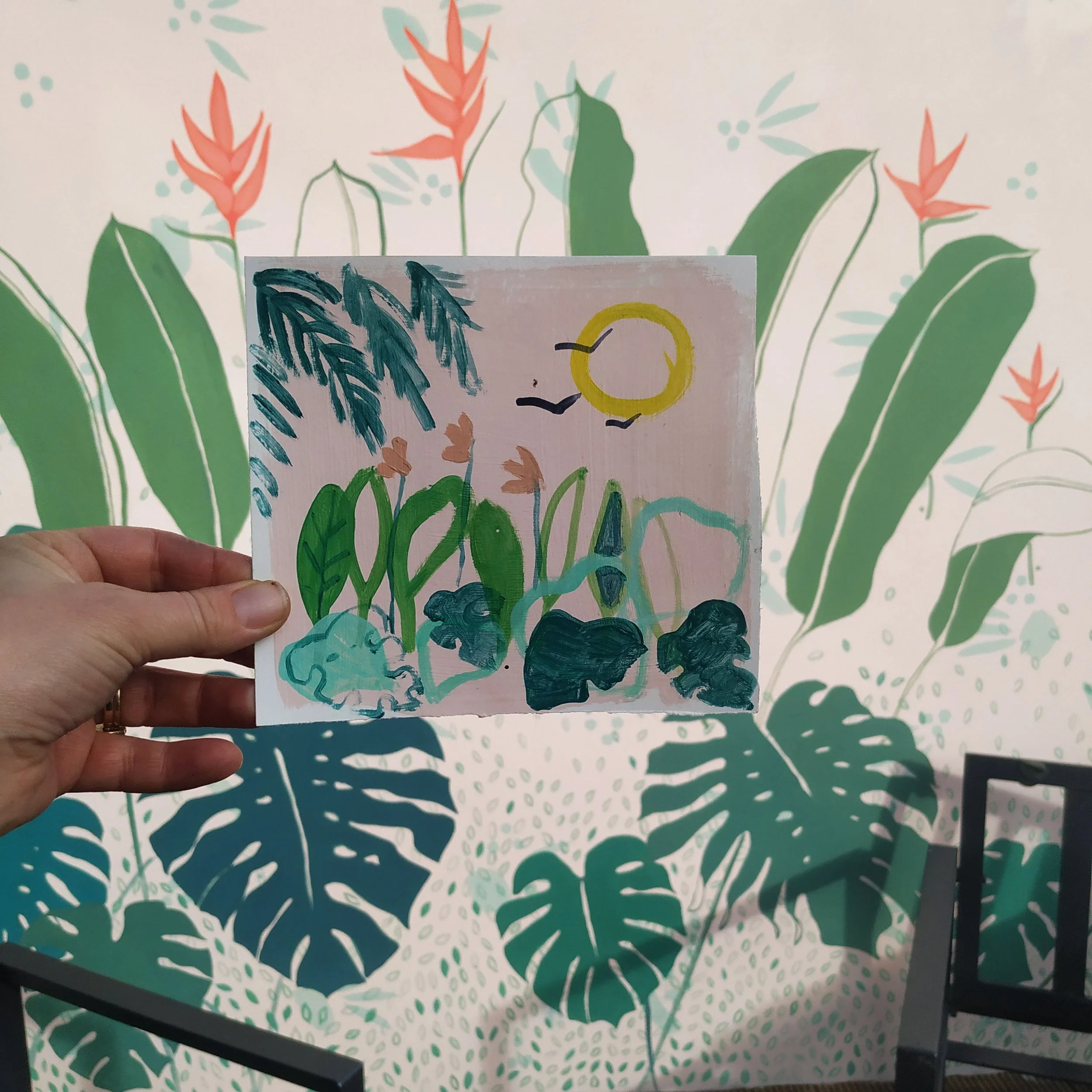Hand holding a colorful painting of a nature scene with plants, flowers, a sun, and birds against a mural of large green leaves and pink flowers.