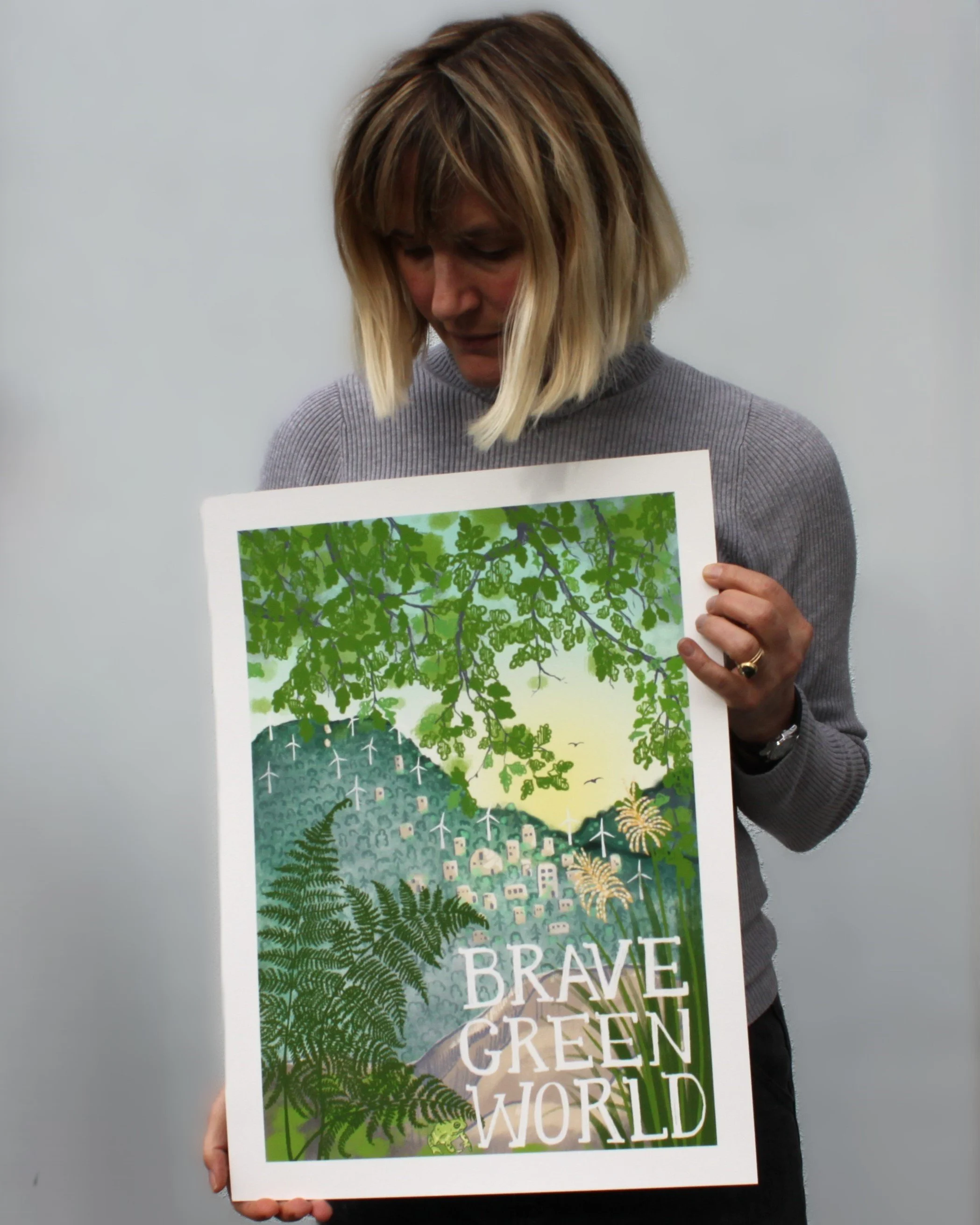 A woman with shoulder-length blonde hair holding a colorful poster featuring a green landscape with trees, wind turbines, a village, and the words 'Brave Green World'.