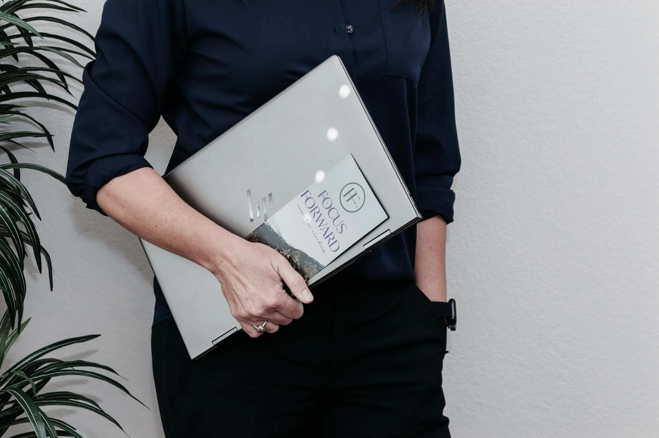 melinda east dressed in a dark blue shirt and black pants holding a silver laptop and a pamphlet titled 'Focus Forward' in front of a white wall, with a plant partially visible on the left.