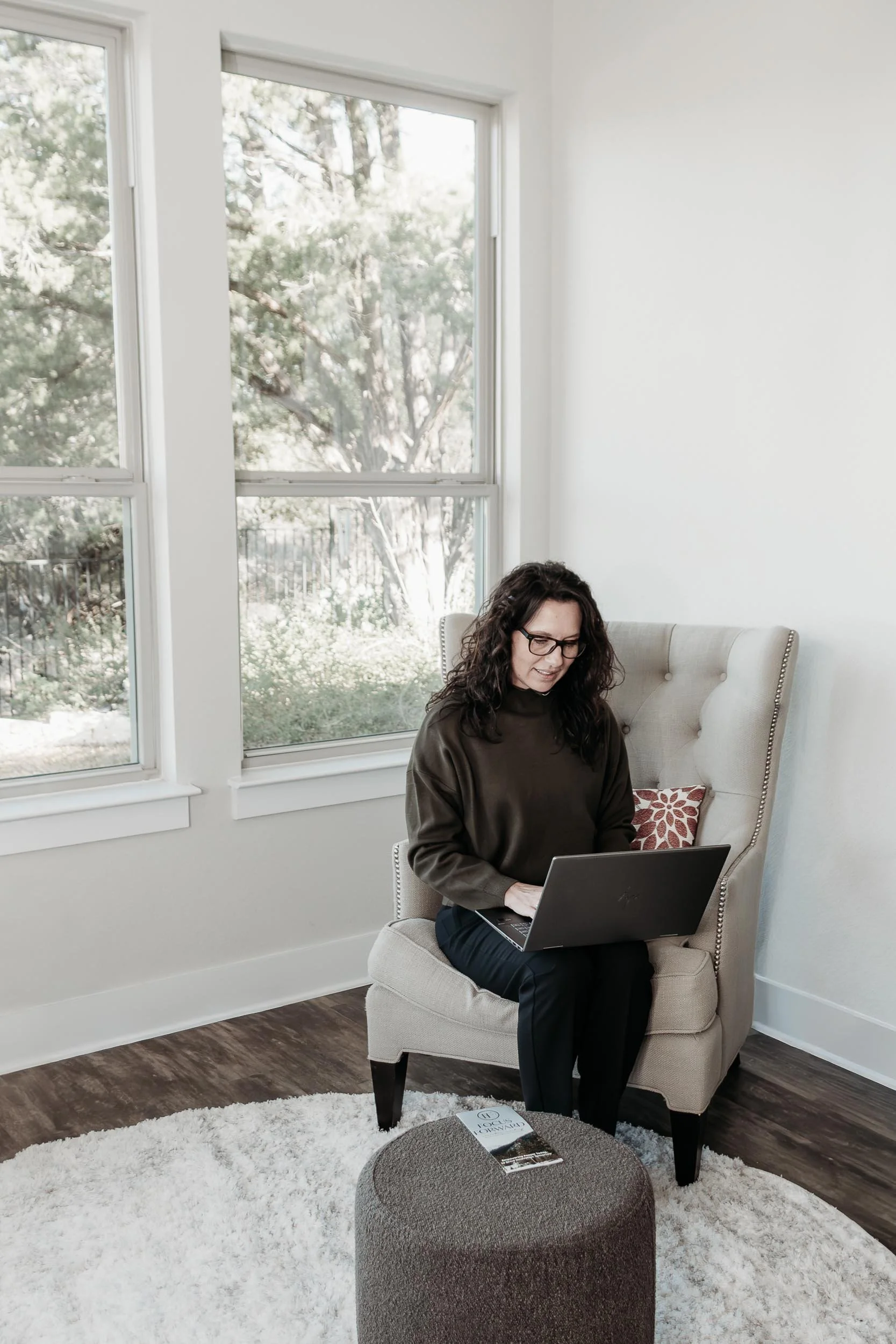 melinda east, working on change management, with curly dark hair and glasses sitting in a beige armchair, working on a laptop, next to a window with a view of trees outside. A round ottoman with a magazine on top is in front of her on a white rug.