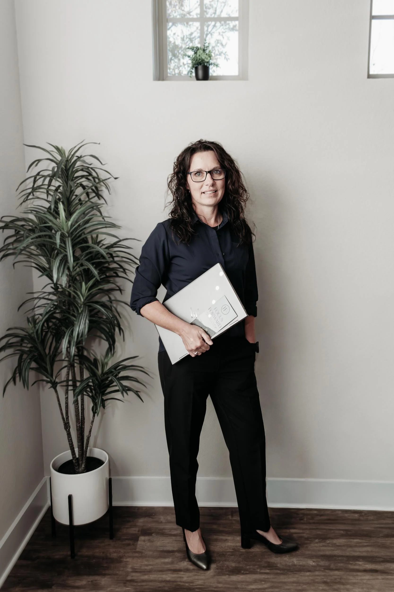 Melinda East with curly dark hair and glasses wearing a black blouse and pants, holding a white folder, stands beside a potted plant, in a room with white walls and small high windows.