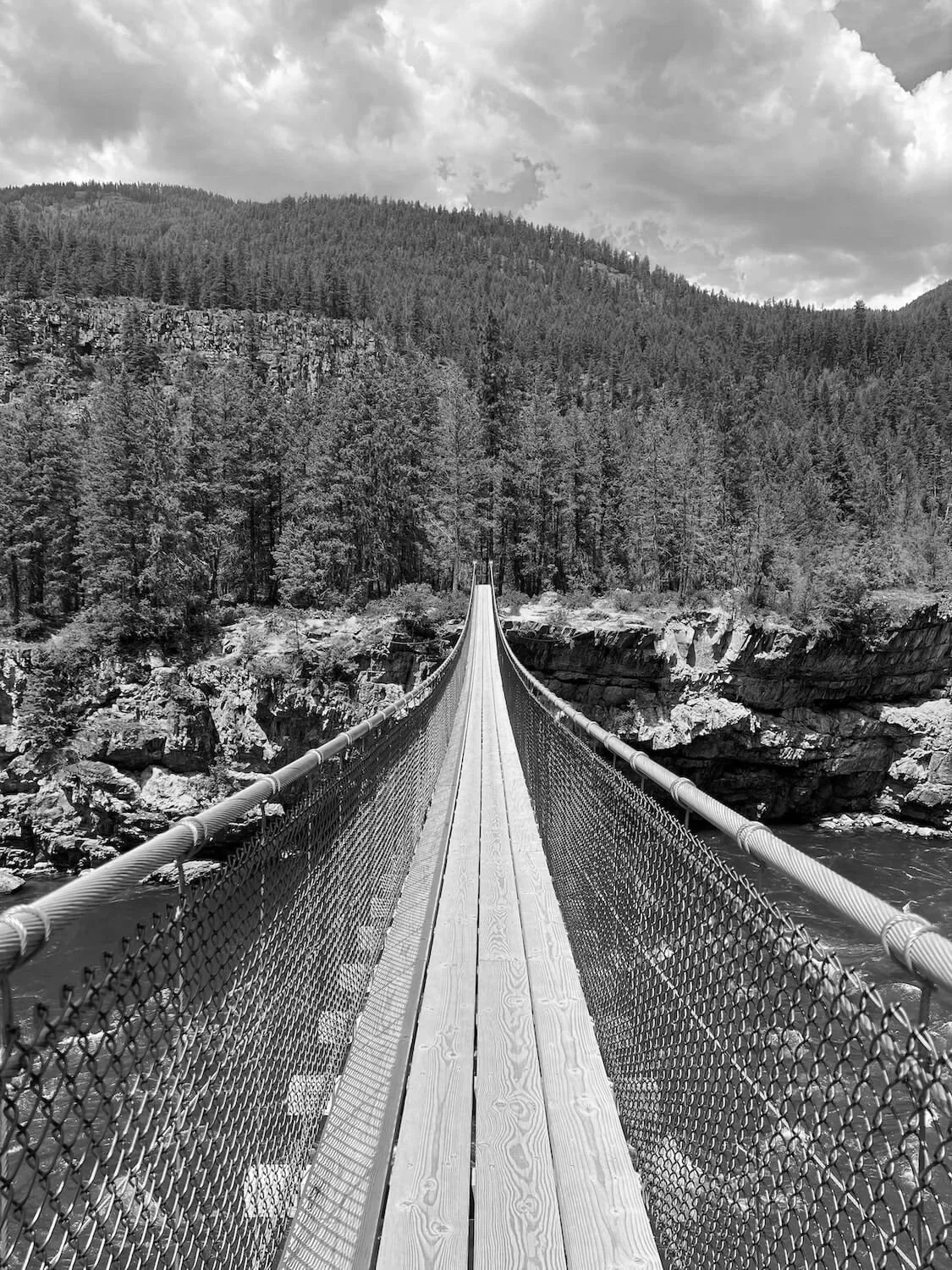 focus forward leadership development. Black-and-white photo of a suspension bridge extending over a river, surrounded by dense forested mountains under a cloudy sky in Montana.