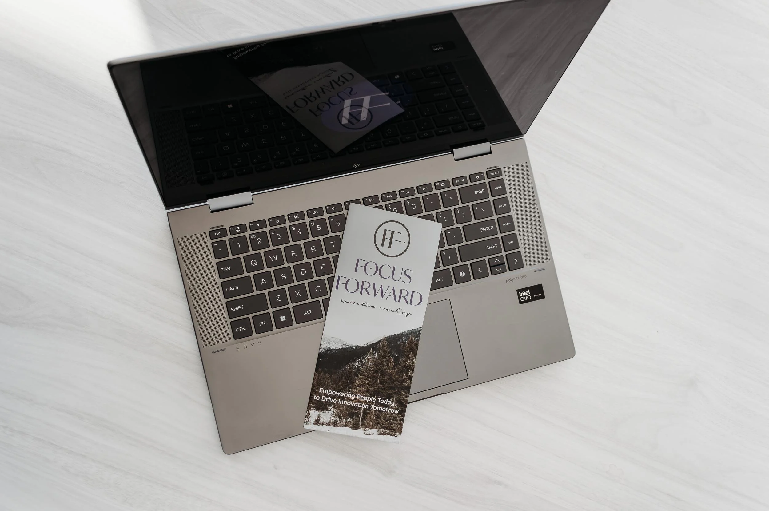 building data culture, A silver laptop with a black keyboard on a white wooden surface, with a brochure labeled 'Focus Forward' resting partially on it.