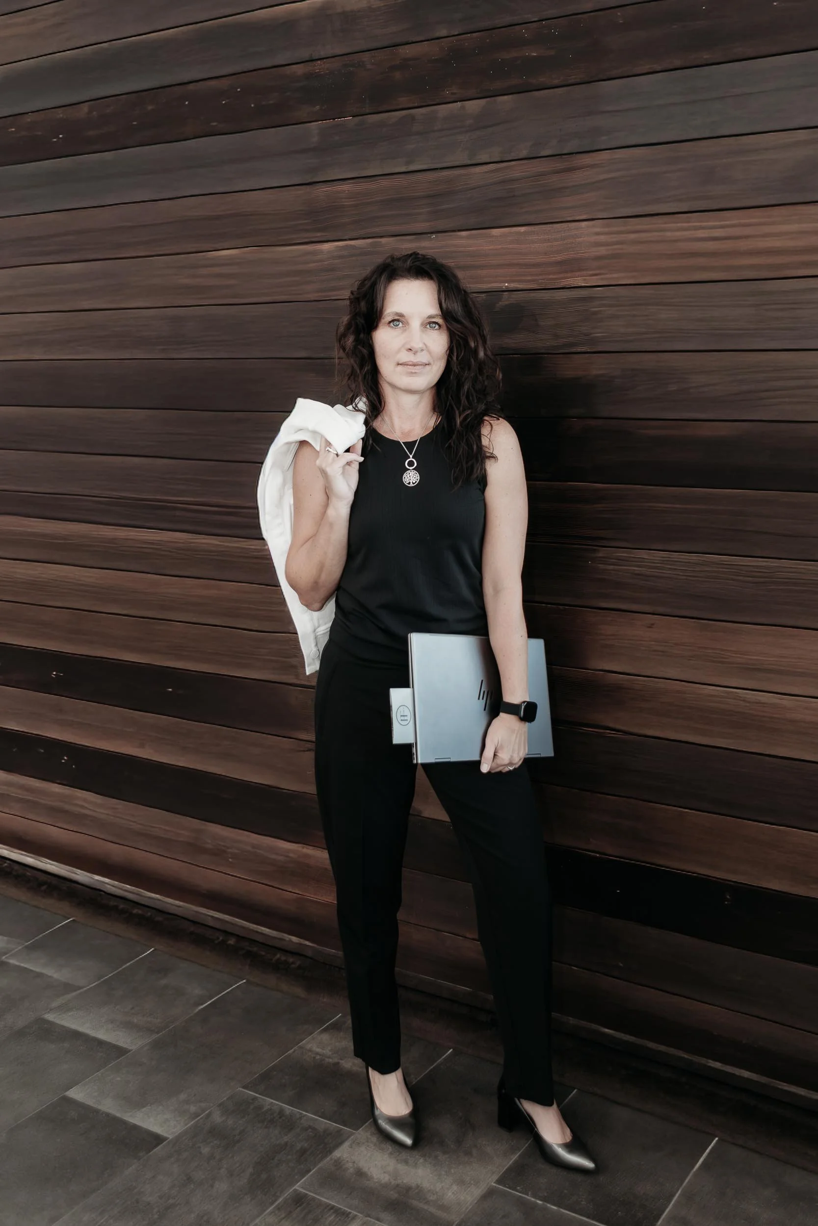 leadership coaching with Melinda East, a woman with dark curly hair dressed in black, wearing a necklace, black pants, and metallic heels, stands against a wooden wall, holding a laptop in one hand and a white jacket slung over her shoulder.