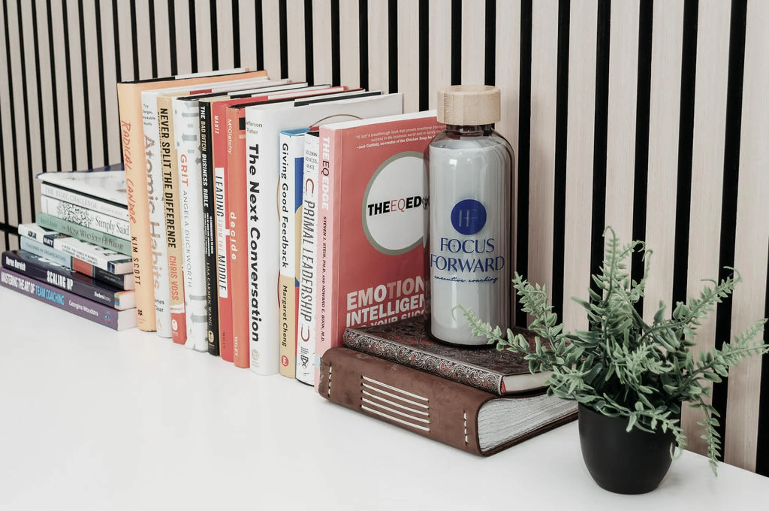 A row of books stacked vertically with a water bottle, a closed notebook, and a potted plant on a white surface against a light wood slat wall.