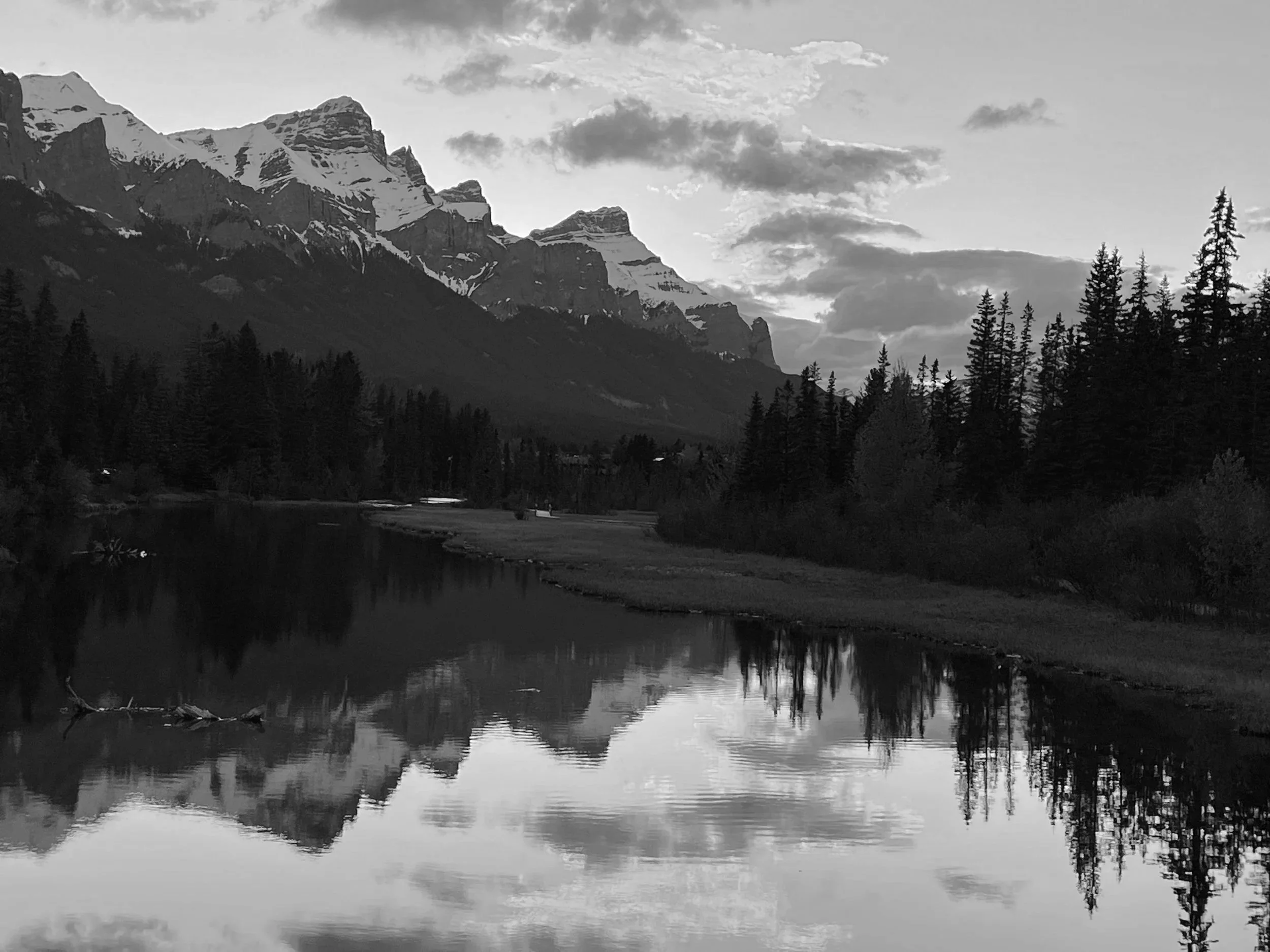advisory services, A black and white photograph of a mountain landscape with snow-capped peaks in the background, a forest of tall trees on the right side, and a calm river reflecting the mountains and sky in the foreground.