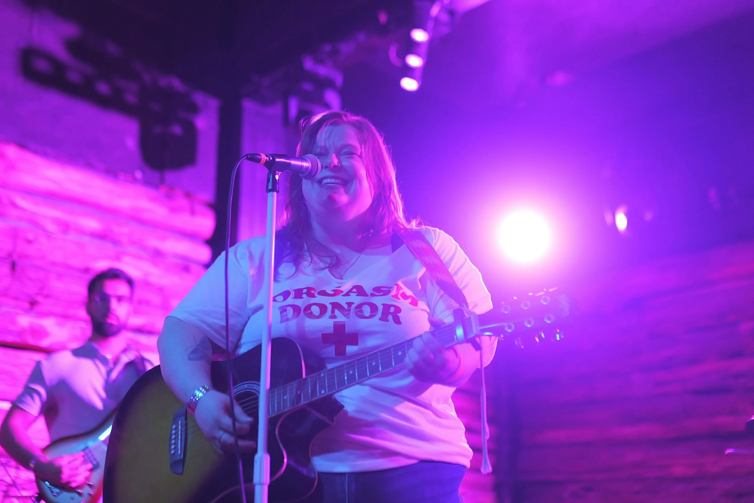 A woman smiling and playing an acoustic guitar on stage with purple and pink lighting, wearing a white T-shirt with text.