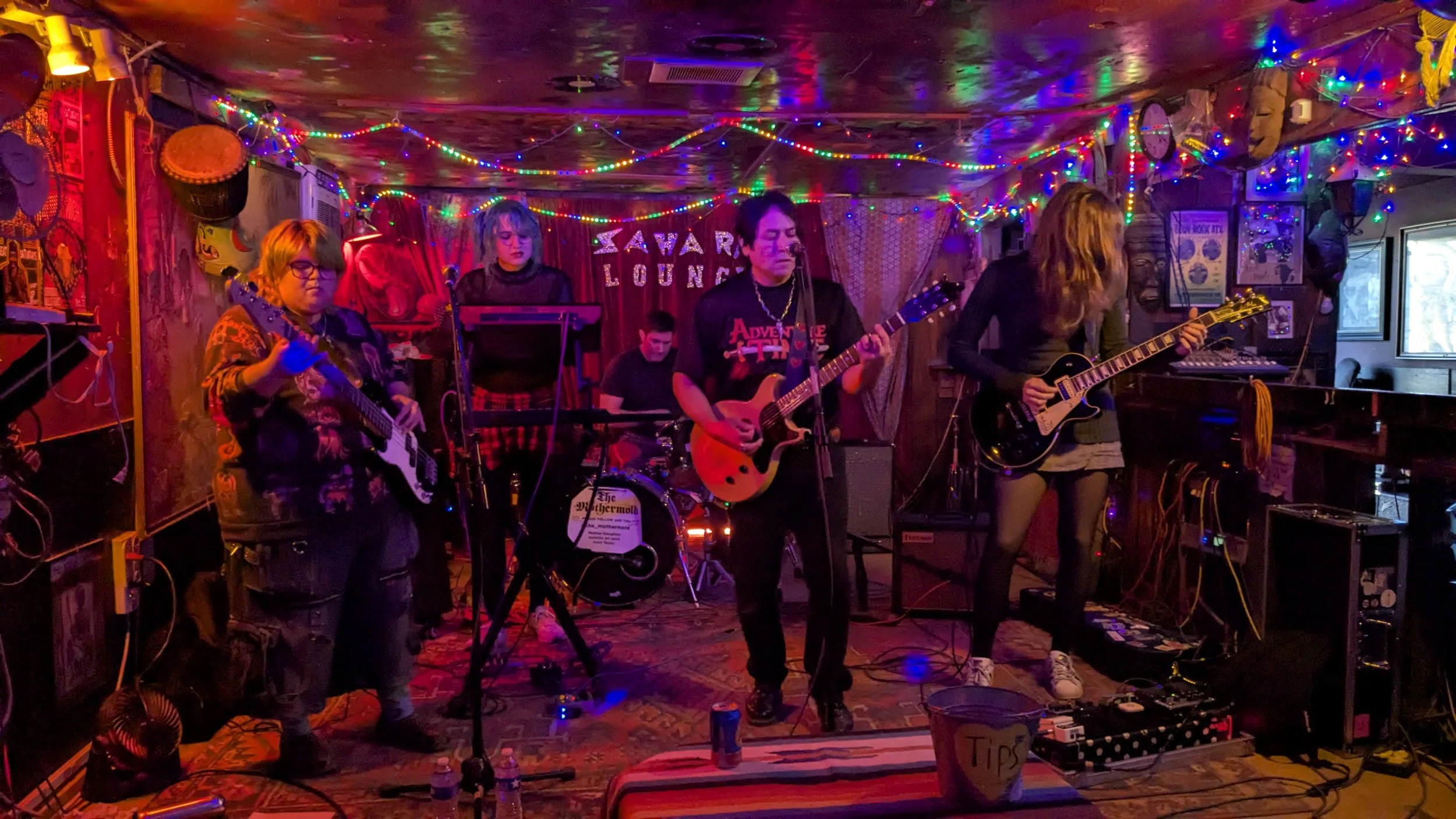 A band performing live on stage at a cozy venue decorated with colorful string lights, with five members playing guitars, drums, and electronics, and a backdrop reading 'SAVAR LOUNGE'.