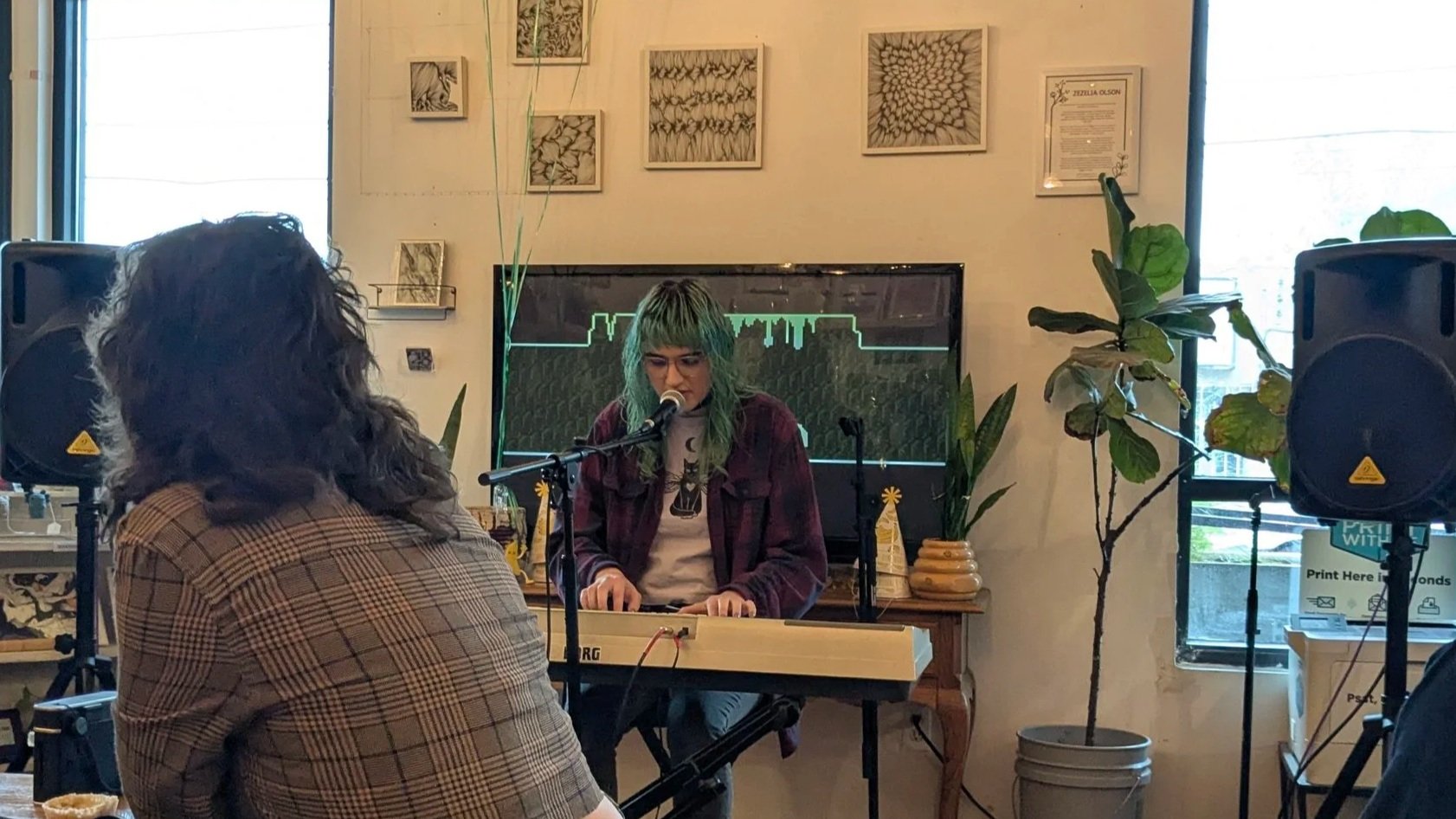 A woman with green hair and glasses playing a keyboard and singing into a microphone indoors, with an audience seated in front of her. The room has framed artwork on the wall, potted plants, and large windows letting in natural light.