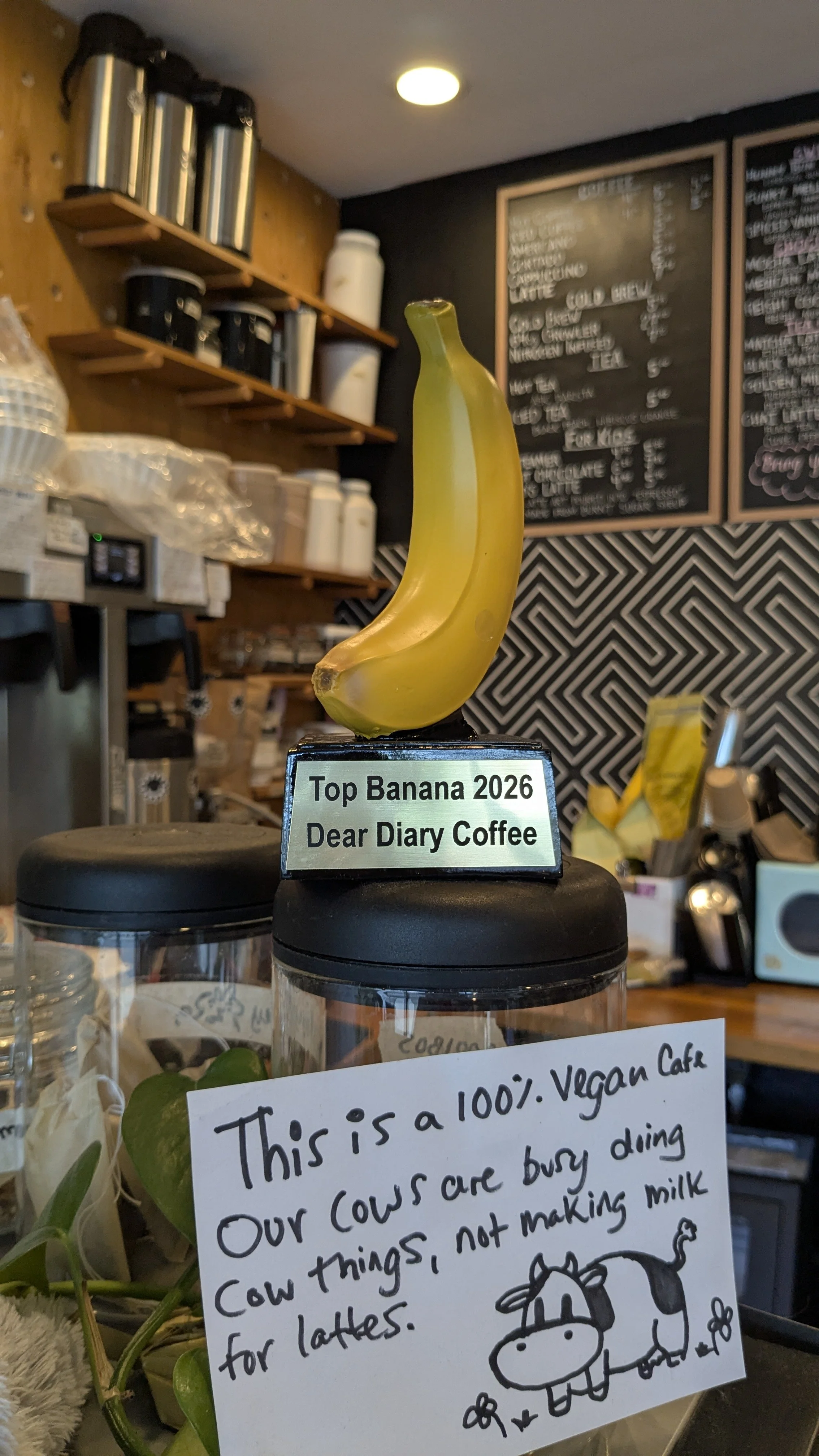 A decorative banana sculpture with a label that reads 'Top Banana 2026 Dear Diary Coffee' in a cafe setting. The background features a chalkboard menu and shelves with various containers.