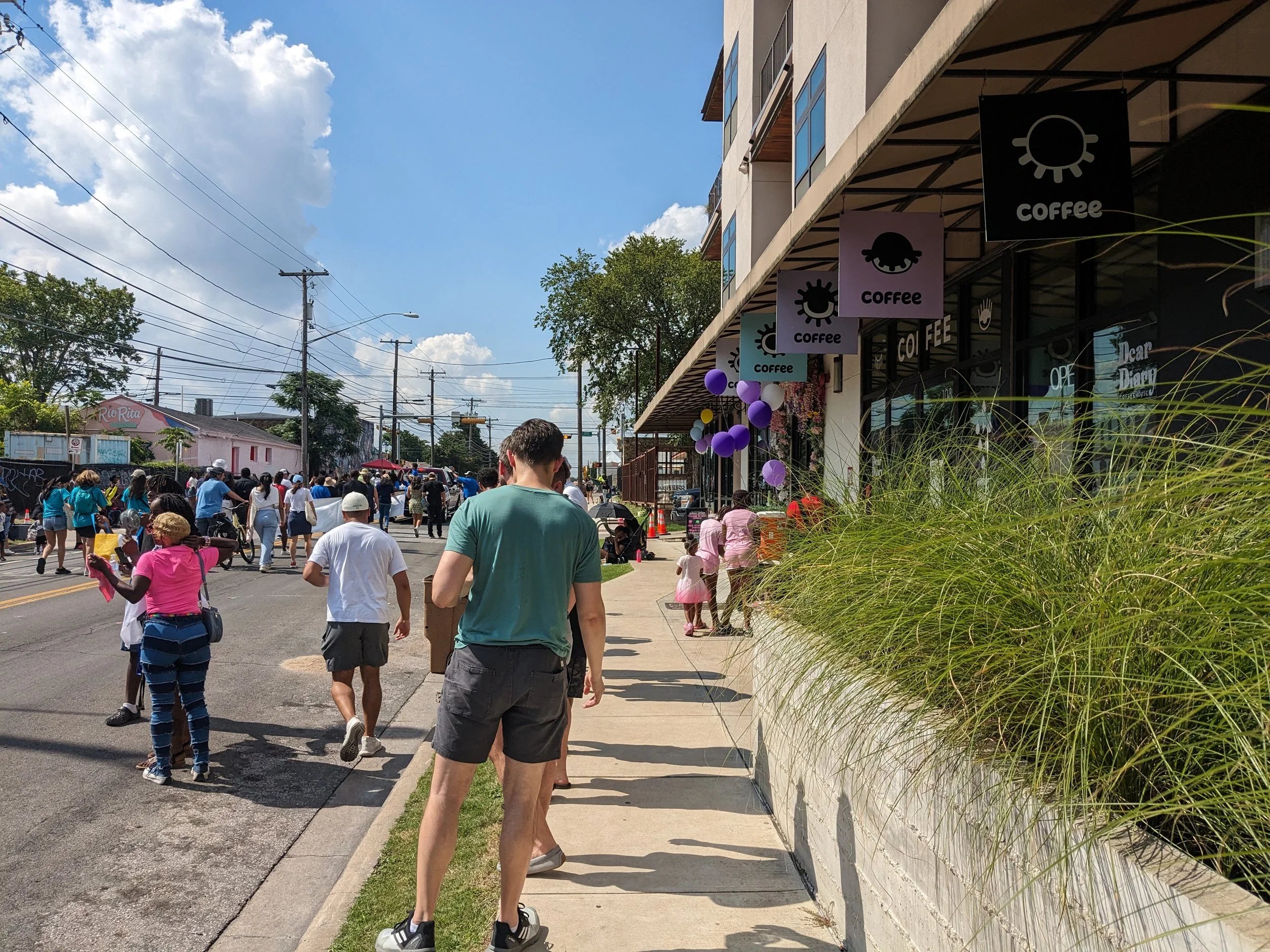 People walking along a sidewalk outside a building with coffee shop signs and balloons. The sky is partly cloudy with power lines overhead.