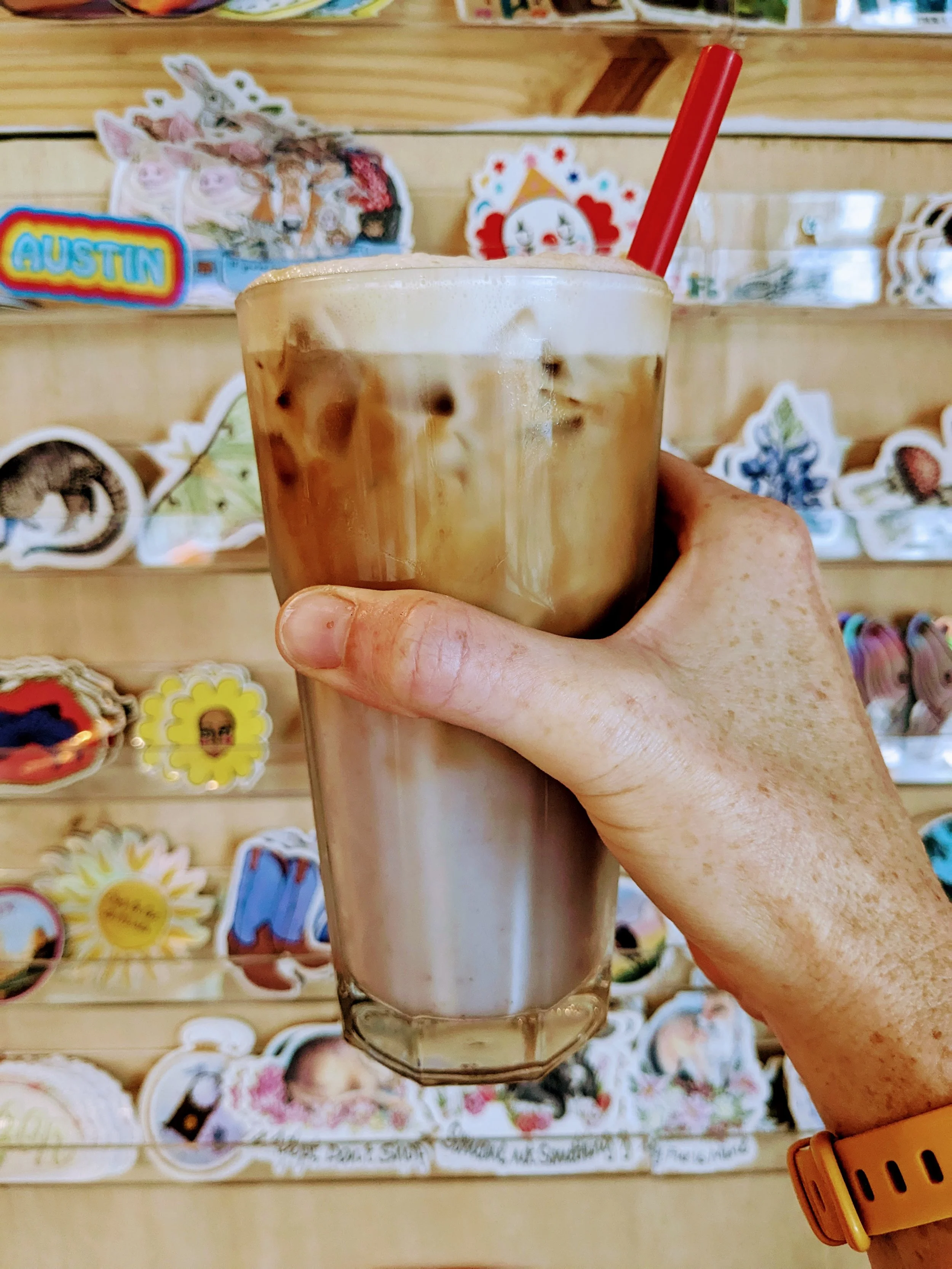 A hand holding a glass of iced coffee with a red straw in front of a wooden display shelf with stickers and decorative items.