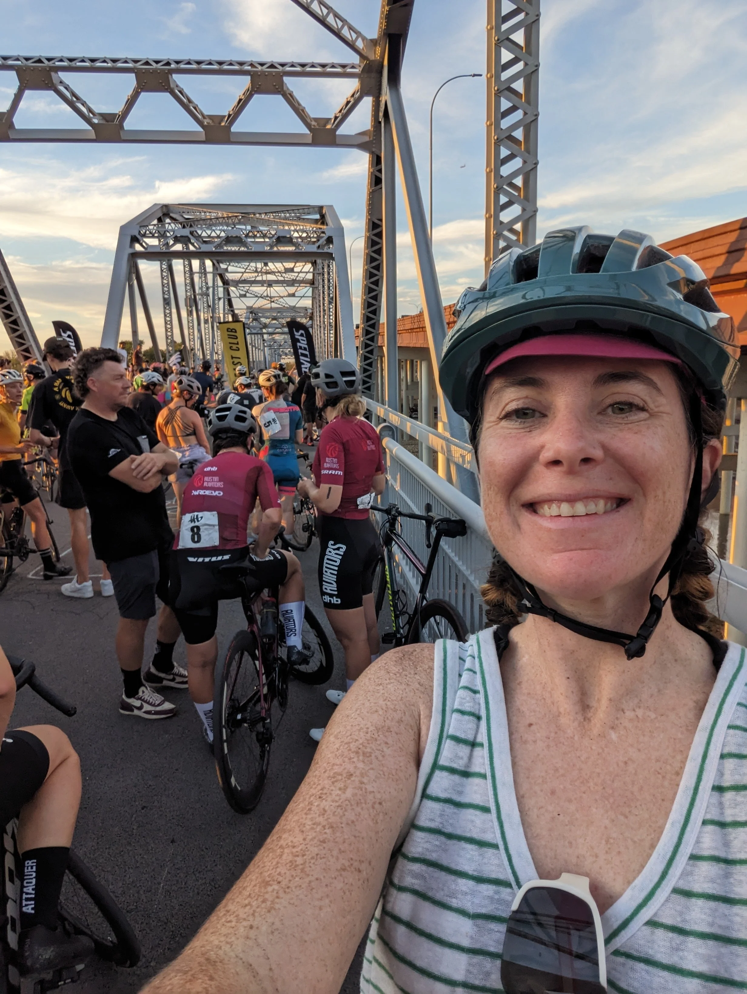 A woman with freckles wearing a bike helmet smiling at the camera, surrounded by other cyclists in a race or event, with a metal bridge and blue sky in the background.