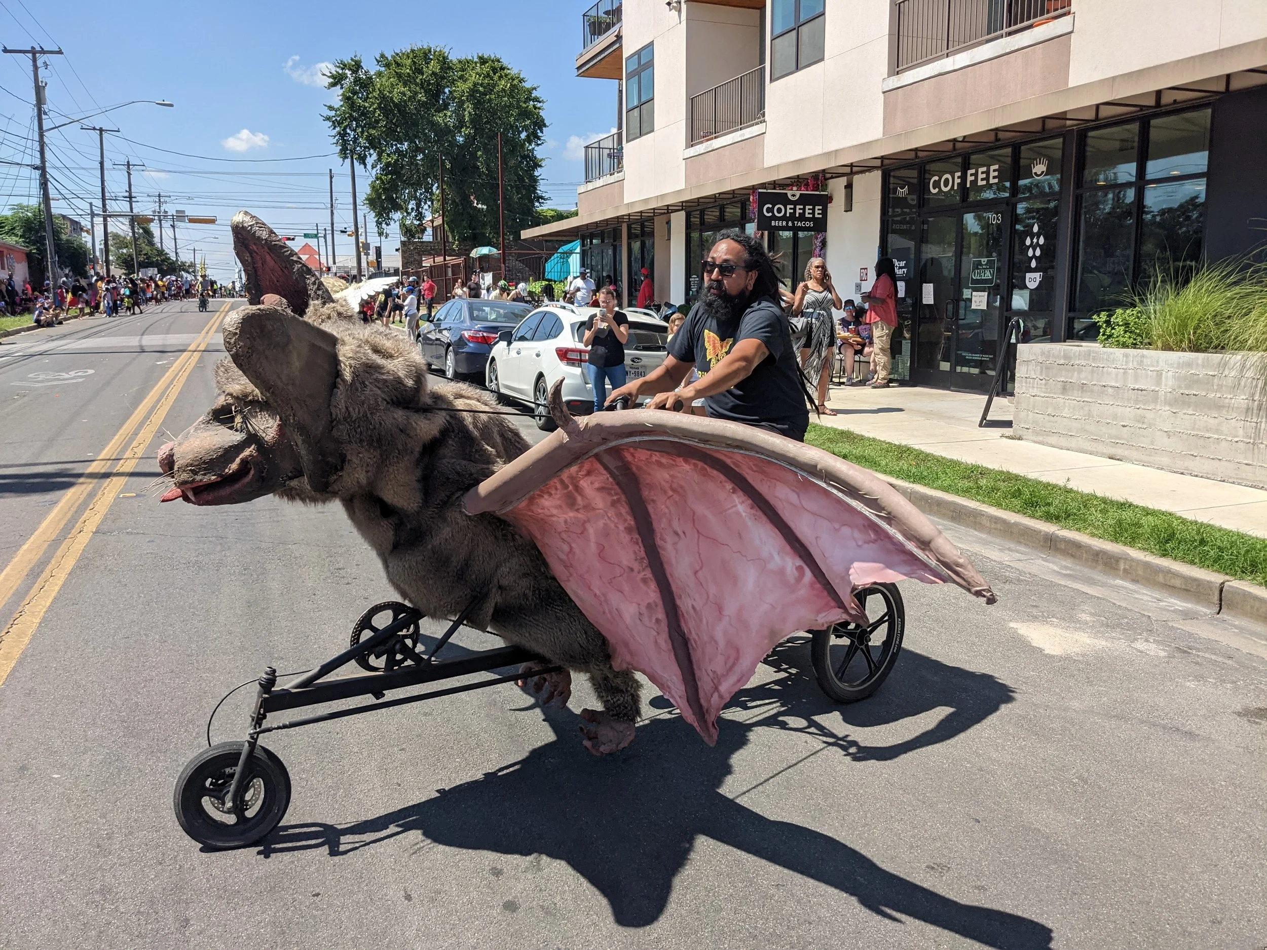 A man with dreadlocks riding a tricycle with a plush kangaroo in a pink pouch on the street during a busy daytime event with people walking and sitting outside a coffee shop.