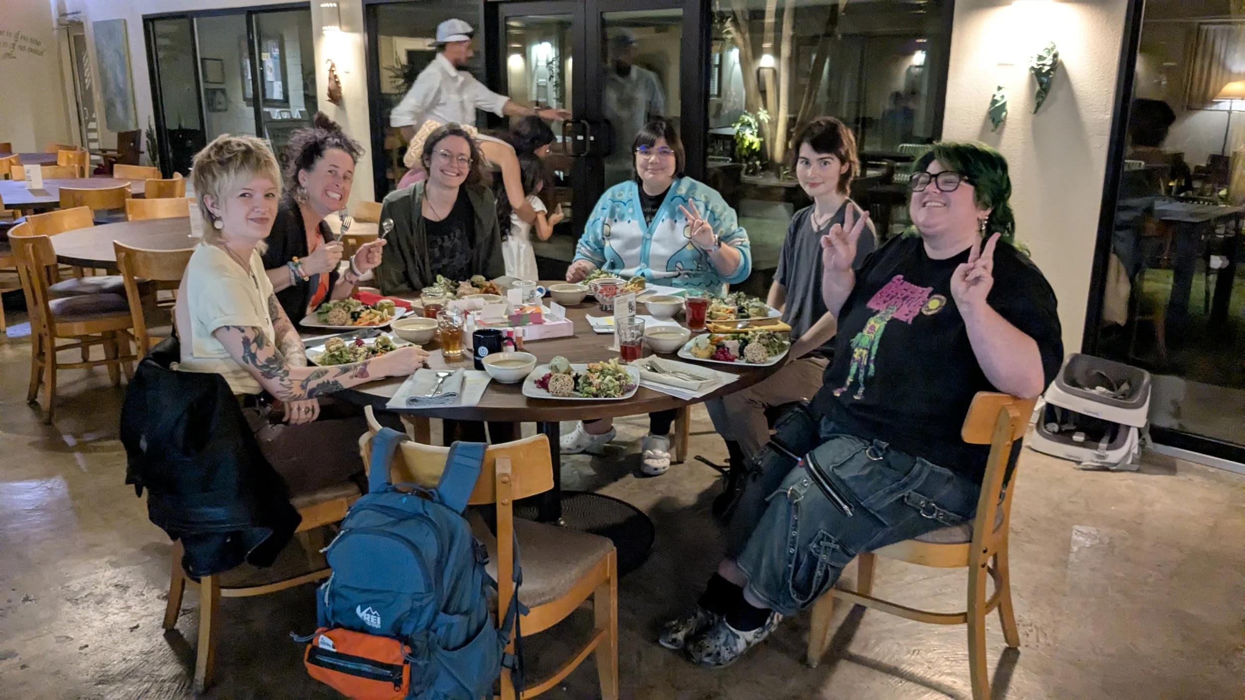 Group of seven friends smiling and making peace signs while sitting at a restaurant table with food and drinks, with some backpacks in the foreground and a waiter in the background.
