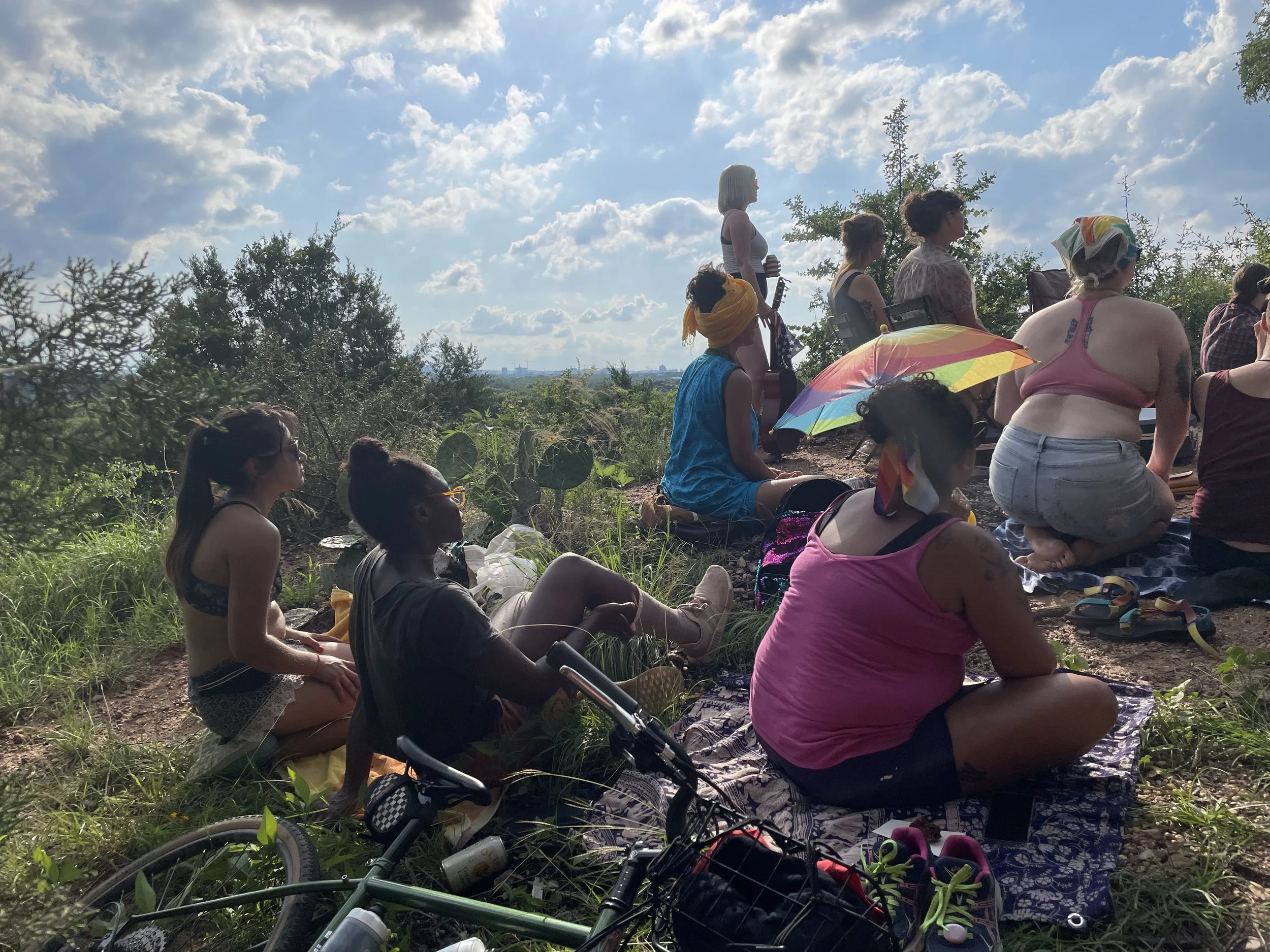 A group of diverse people sitting and standing outdoors on a grassy area with trees and bushes, some holding umbrellas and musical instruments, under a partly cloudy sky on a sunny day.