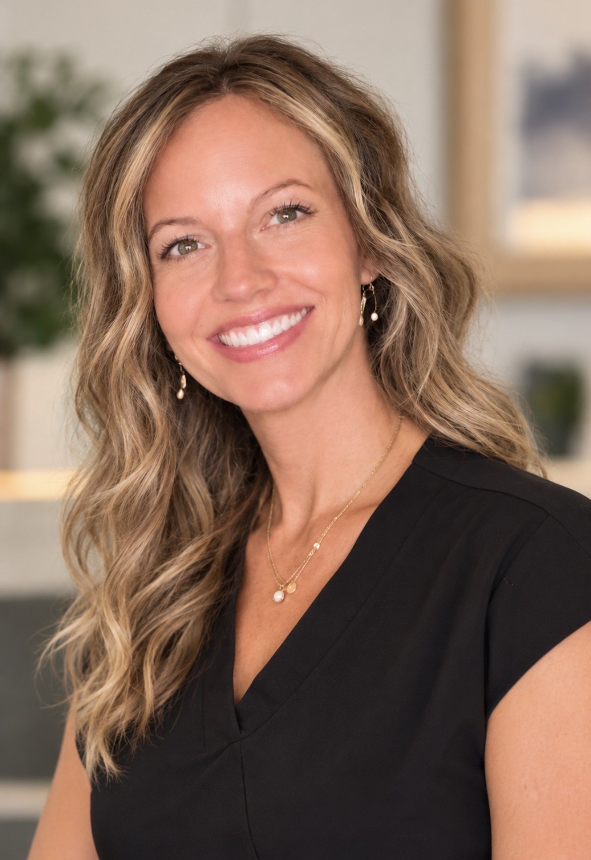 A professional woman with long, wavy blonde hair smiling at the camera, wearing a black top, jewelry, and earrings, in an indoor setting with blurred background.