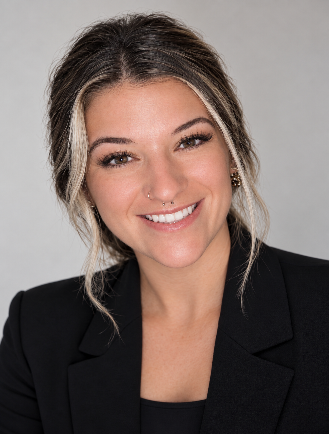 Close-up portrait of a smiling woman with short, wavy hair and multiple facial piercings, wearing a black blazer.